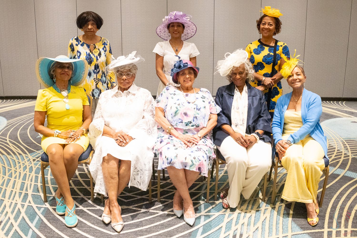 Eight women in colorful dresses and decorative hats posing for a group photo at a formal event.