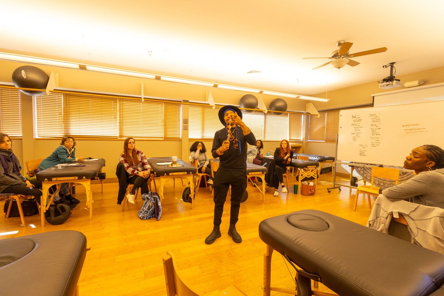 Instructor in black hat speaking to students seated on massage tables in a wellness classroom setting.