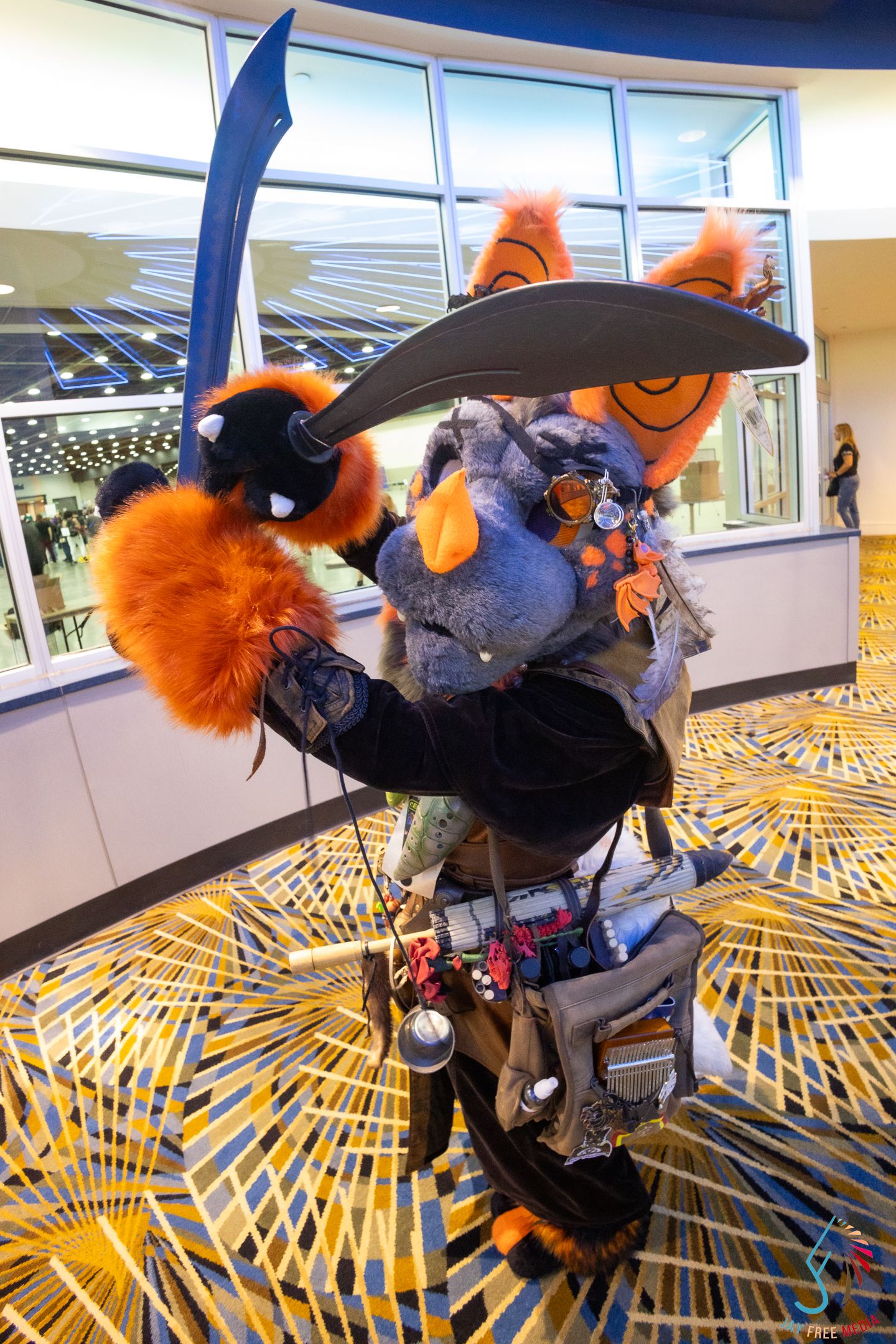 Furry convention cosplayer in orange and gray wolf suit wielding a large sword prop at an indoor event.