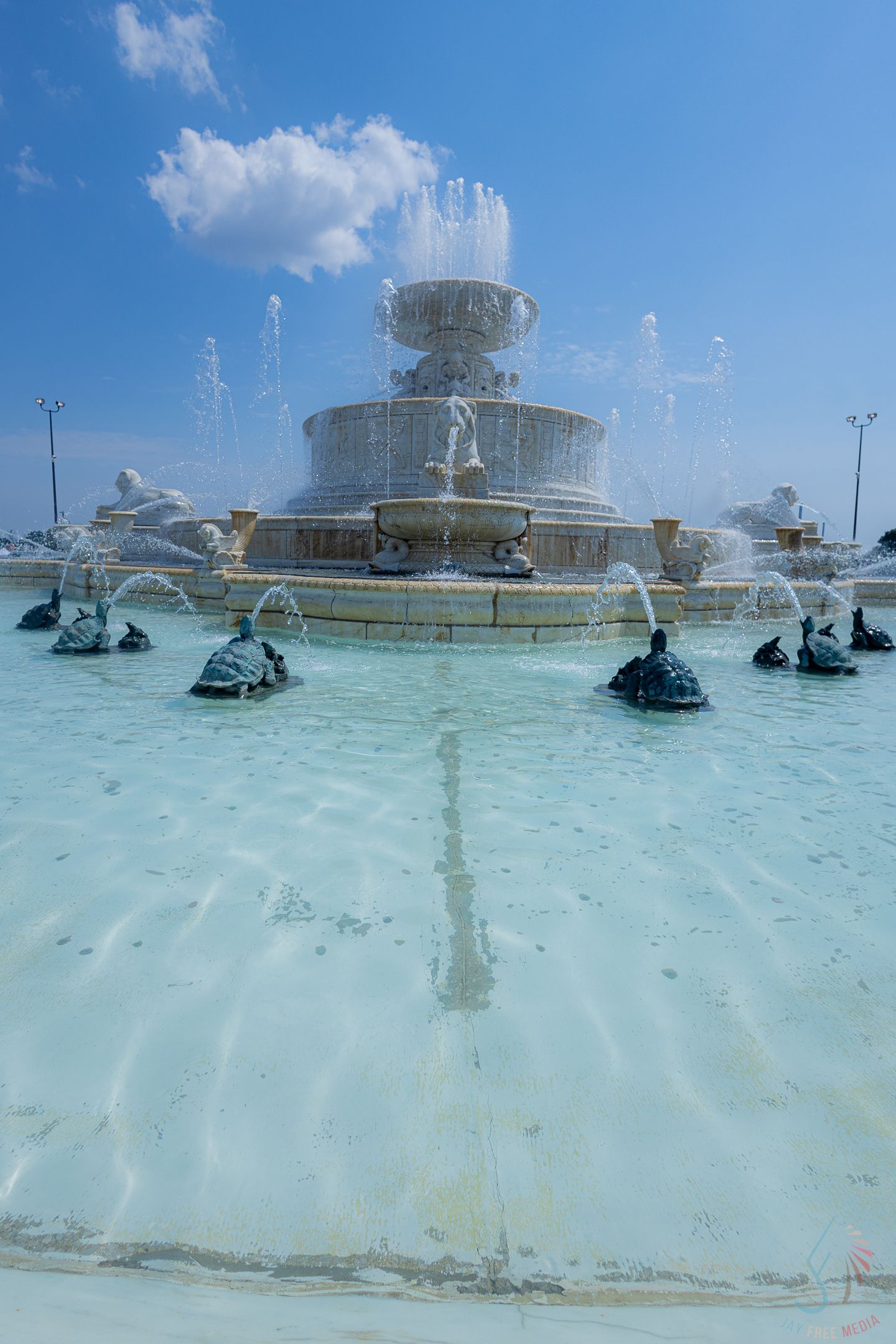 Grand tiered white stone fountain with bronze turtle sculptures in a clear blue pool under a bright sky.