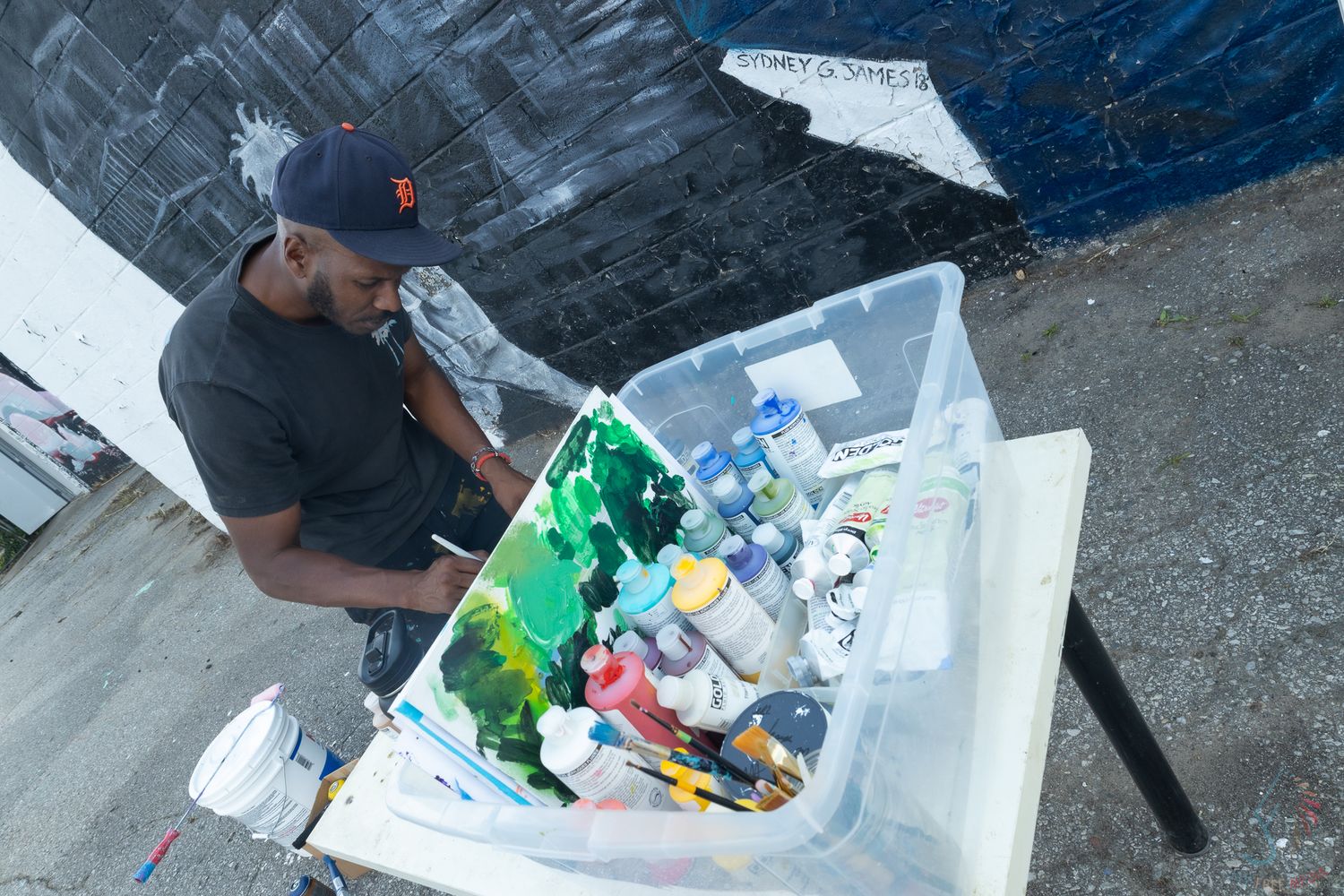 Artist mixing paints in a plastic container outdoors near a large mural in progress.