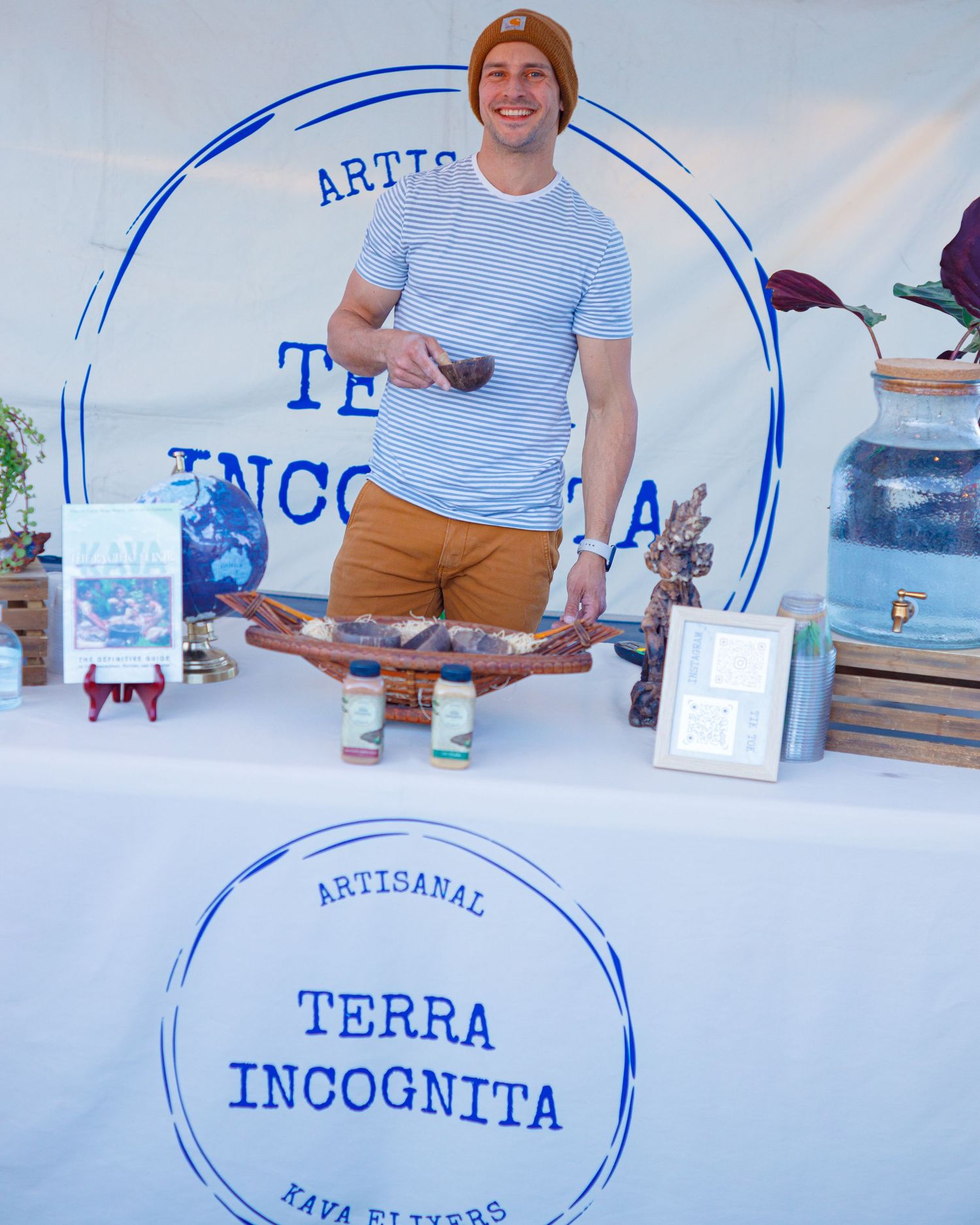 Smiling man standing behind Terra Incognita artisanal kava elixirs vendor table at market.