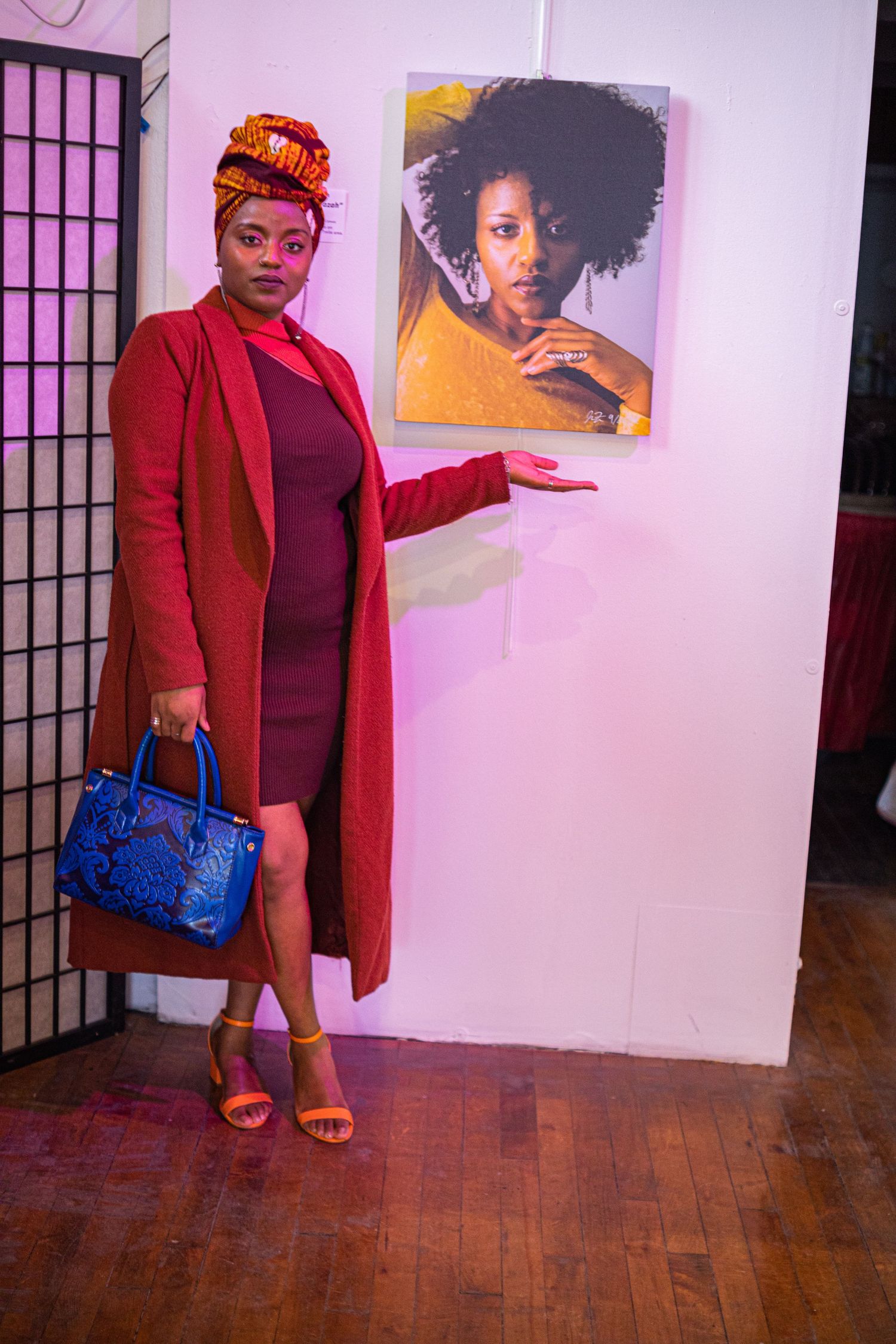 Elegant woman in red coat and colorful headwrap poses beside a portrait photograph at an art gallery.
