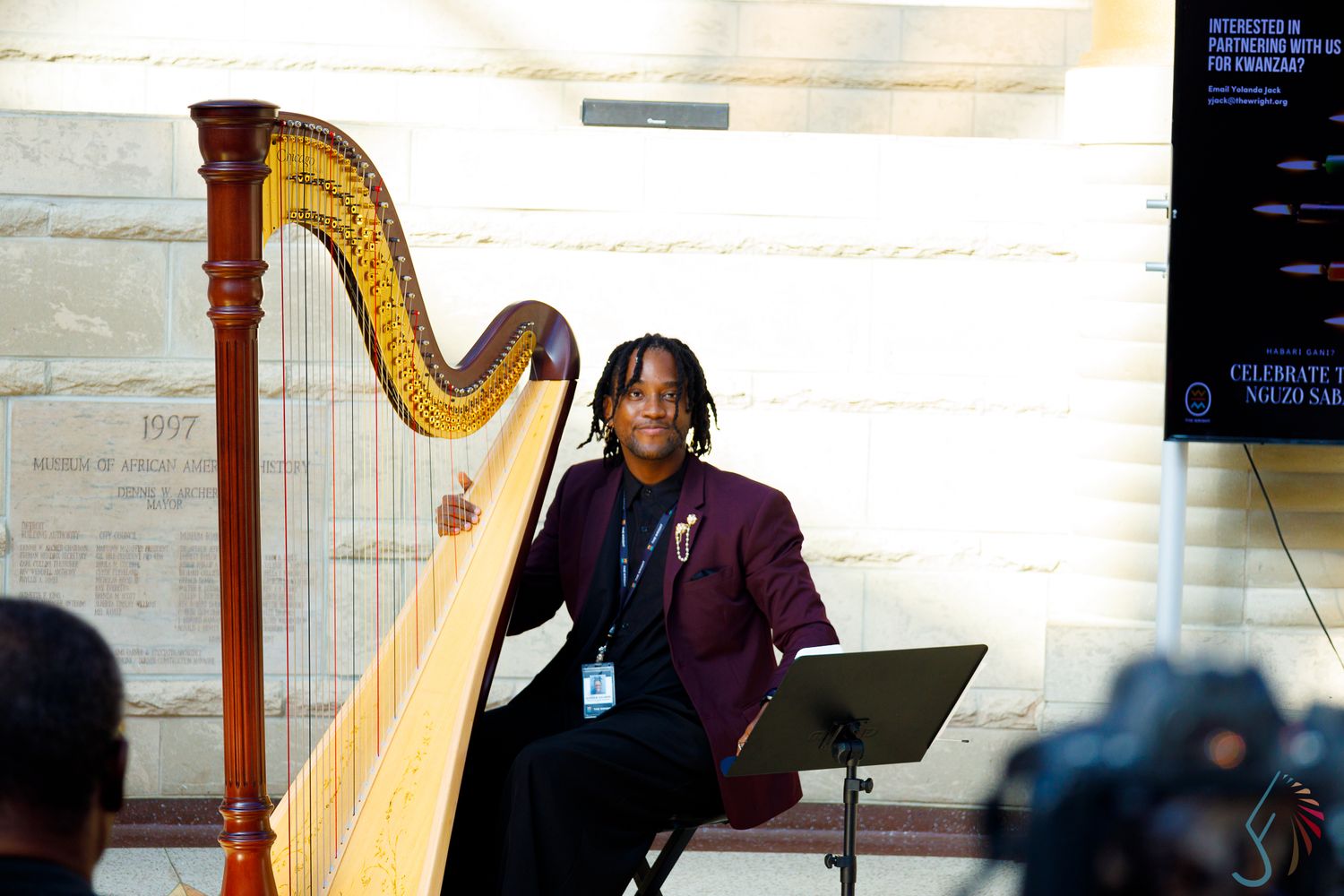 A smiling harpist in a burgundy blazer performs outdoors near a golden harp on a stage with a music stand.