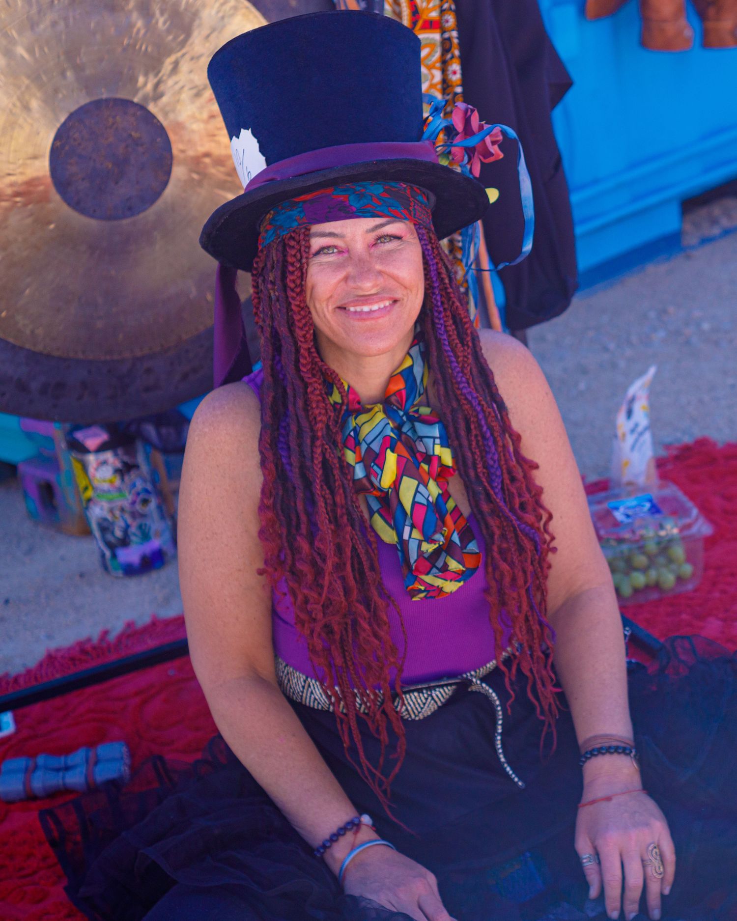 Woman with colorful dreadlocks and top hat sitting near gong at outdoor festival.