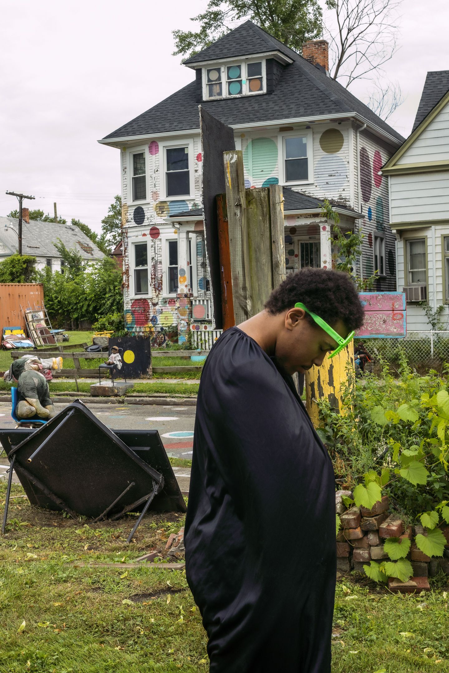 Young man with green glasses stands in front of a colorful decorated house in an urban neighborhood.