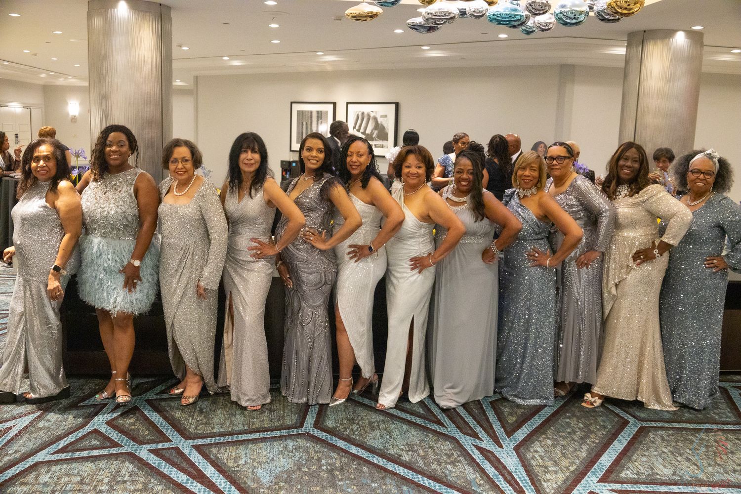 Group of women in elegant silver gowns posing together at a formal event in a decorated ballroom.