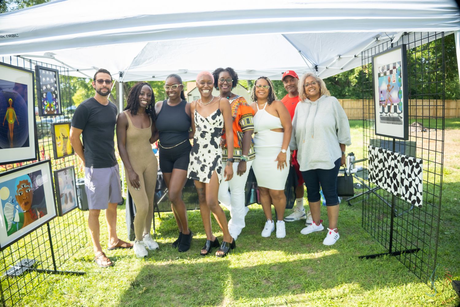 Group of friends posing together under a white tent at an outdoor art festival on a sunny day.