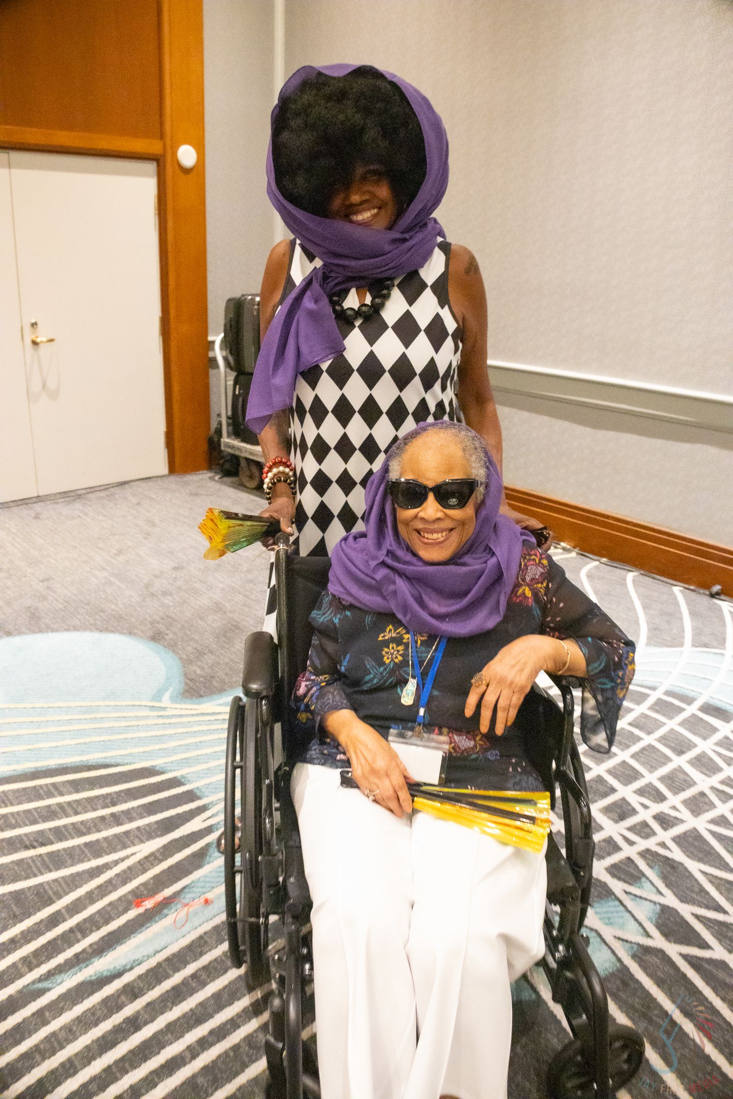 Two women in purple scarves, one standing behind the other seated in a wheelchair, smiling at an indoor event.