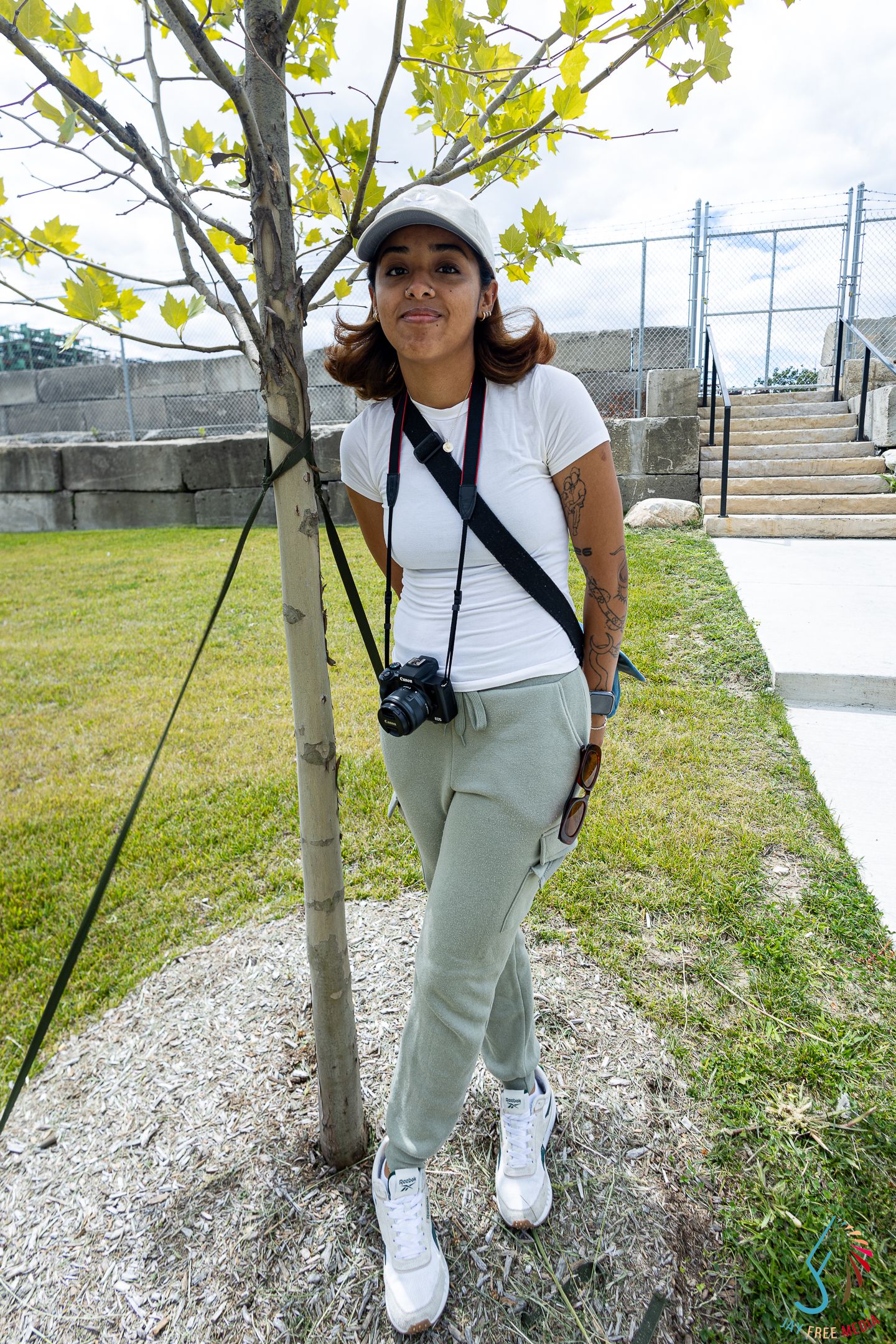 Young woman in white tee, sage green joggers, and cap poses by a tree outdoors with a camera around her neck.