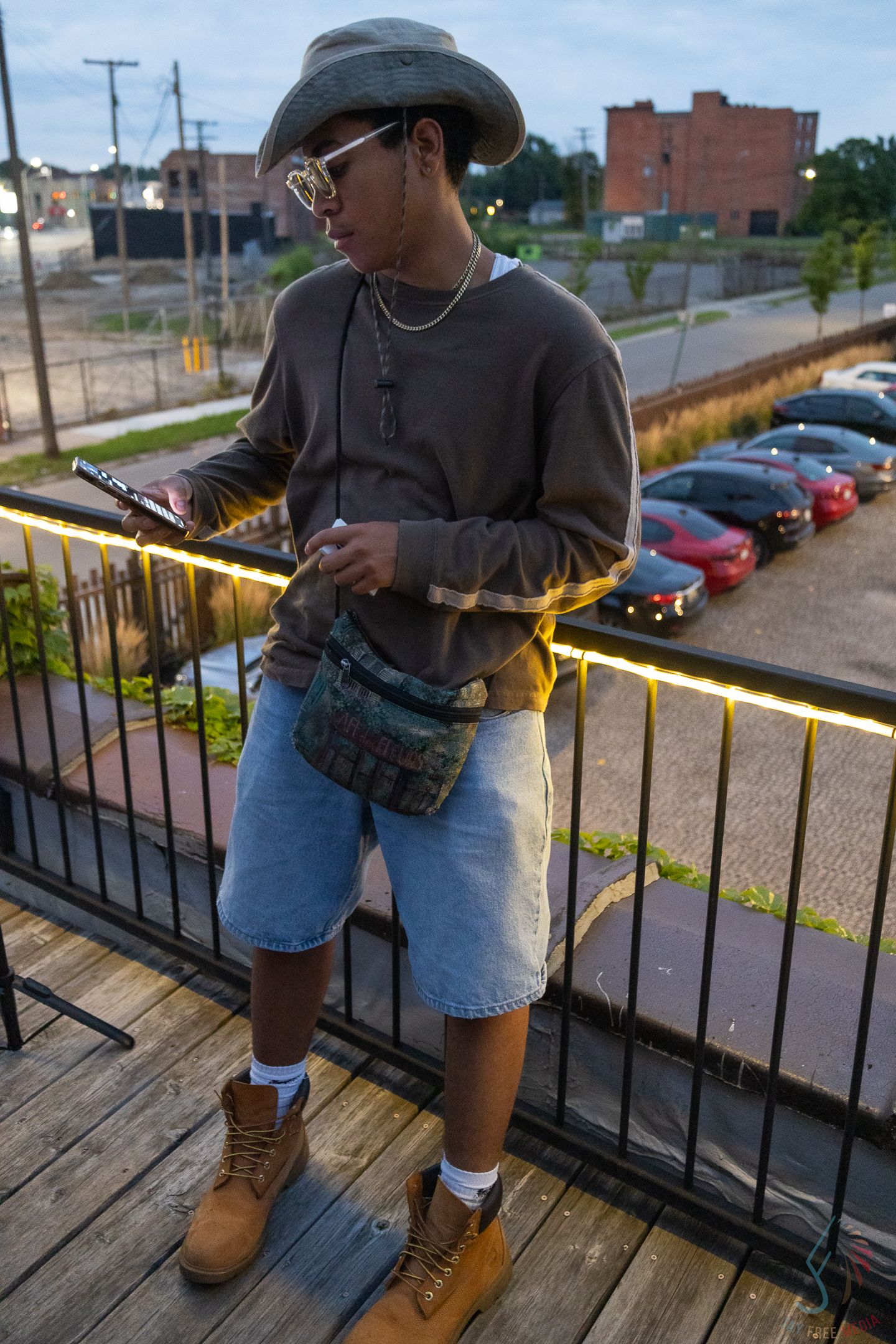 Stylish young man in bucket hat and denim shorts using phone on illuminated outdoor deck.