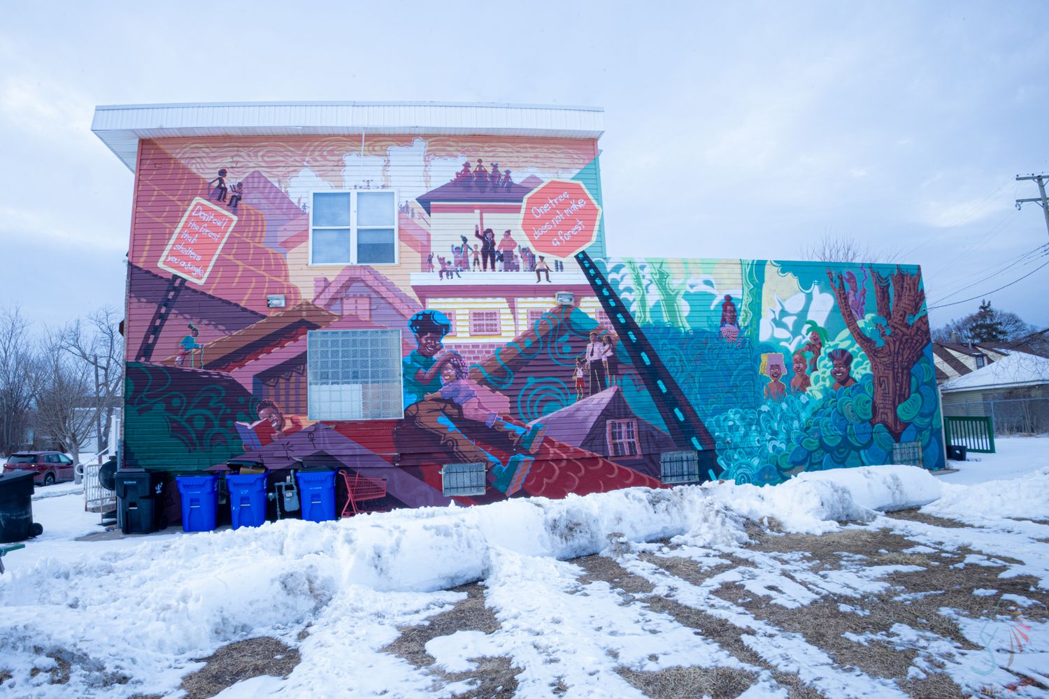Colorful urban mural on brick building exterior in winter, depicting vibrant community scenes with snow on ground.