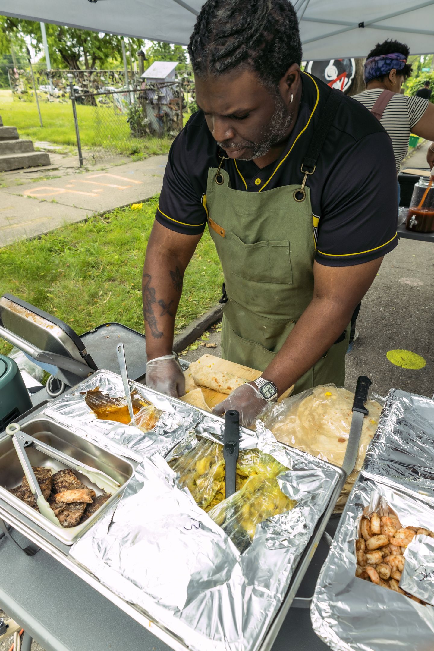 A man in a green apron serves food from aluminum trays at an outdoor food stall during a community event.