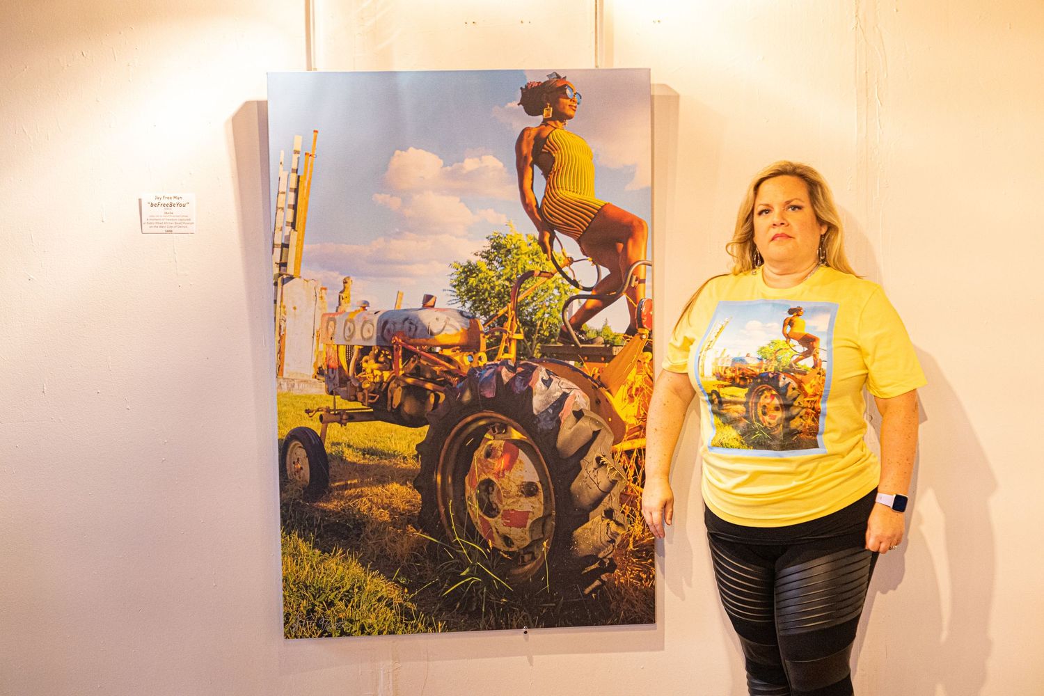 Woman in yellow t-shirt stands beside large vibrant artwork of figure on tractor at gallery.