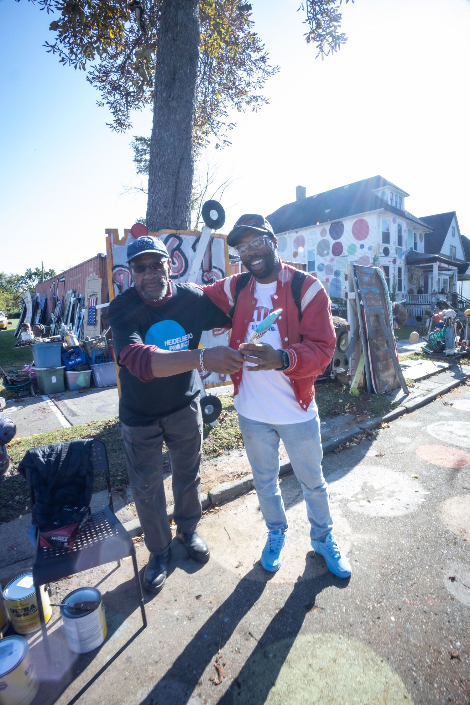 Two men smile and pose together outdoors near an urban art installation on a sunny day.