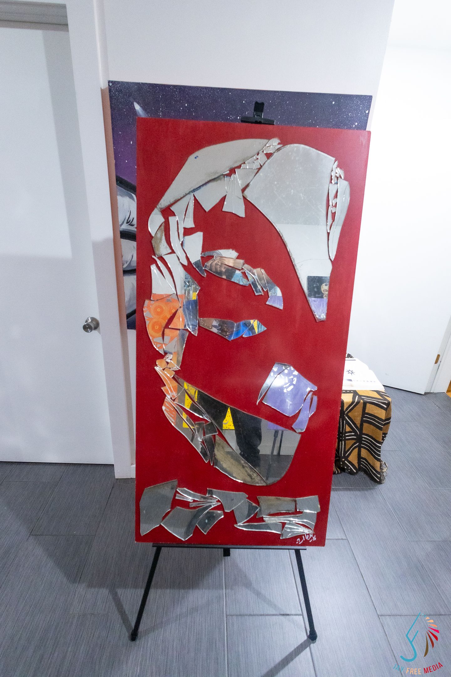 Red canvas artwork with shattered mirror fragments arranged in abstract design, displayed on black easel in white gallery space.