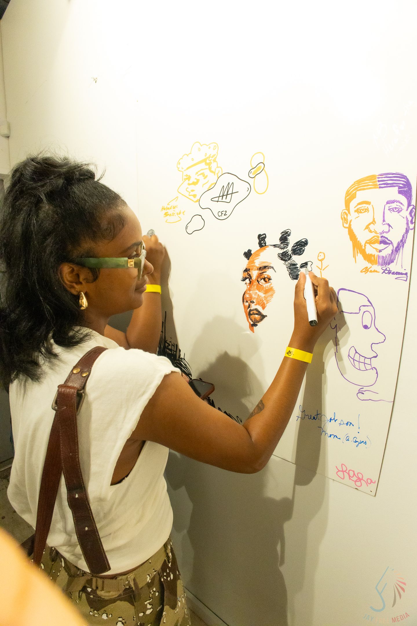 Young Black woman with glasses draws colorful portraits on a white wall at an art event.