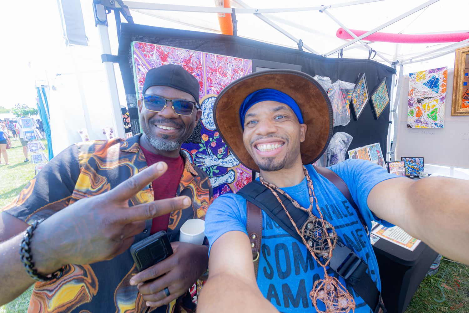 Two men smile for a selfie at an outdoor art festival booth displaying colorful artwork.