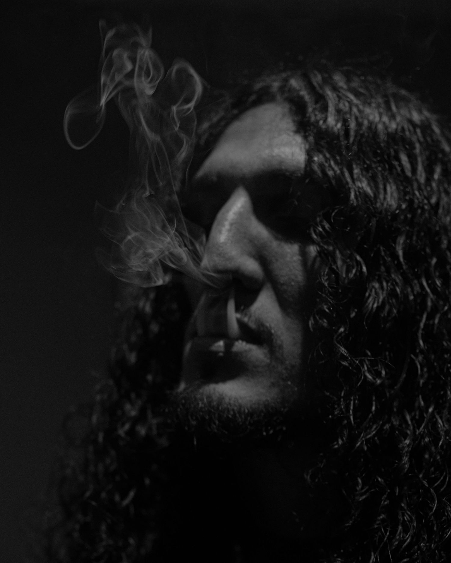 Black and white portrait of long-haired man exhaling smoke in dramatic low-key lighting.