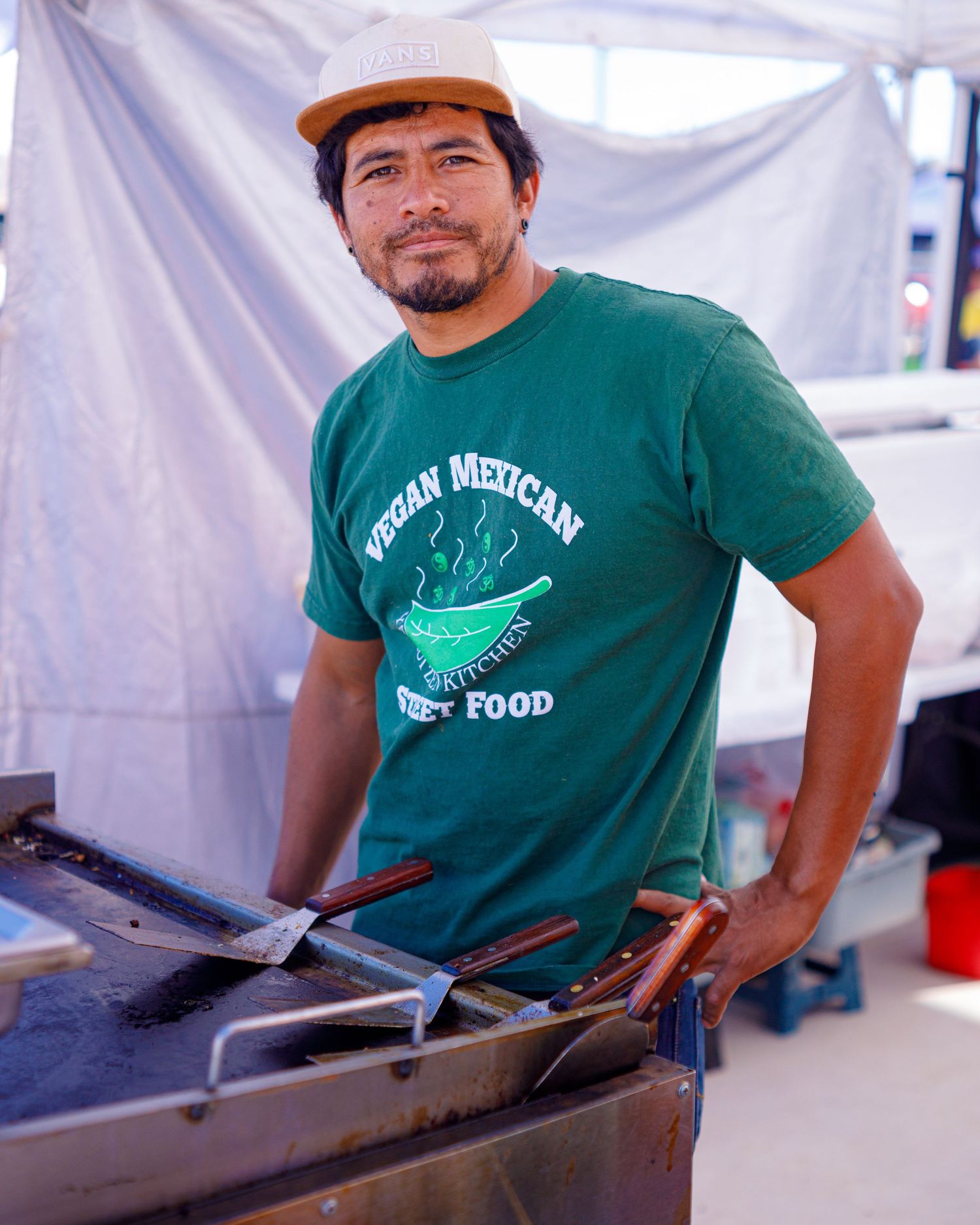 Man in green Vegan Mexican street food shirt cooking at outdoor festival grill.
