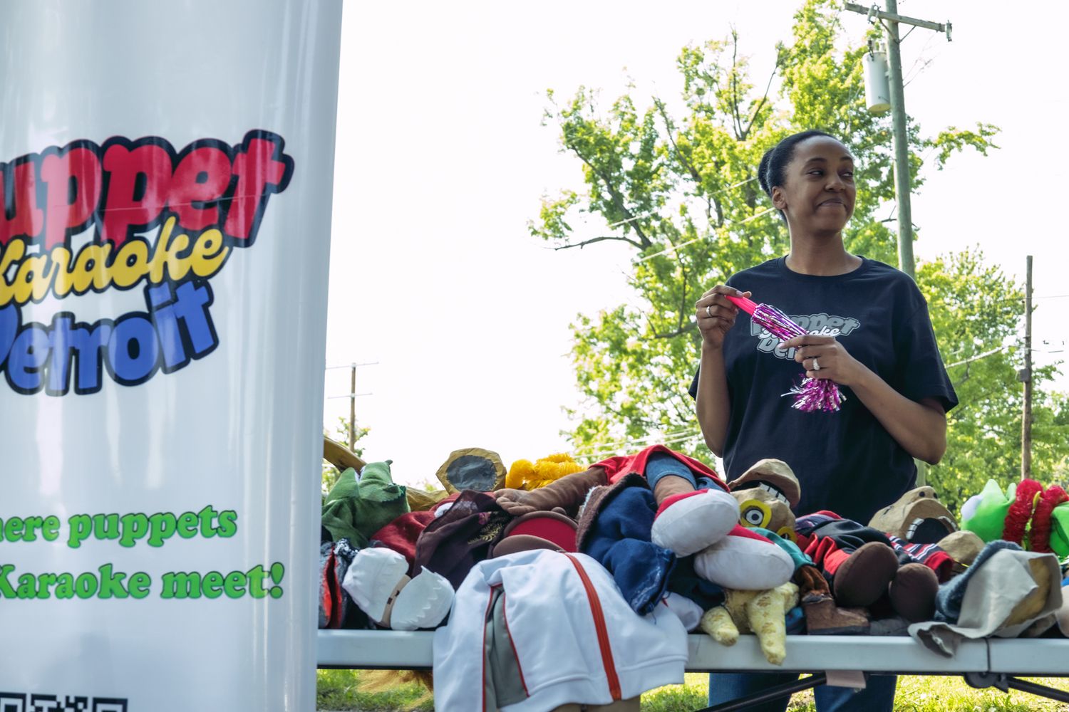 A woman stands behind a pile of colorful puppets at the Puppet Karaoke Detroit outdoor event booth.