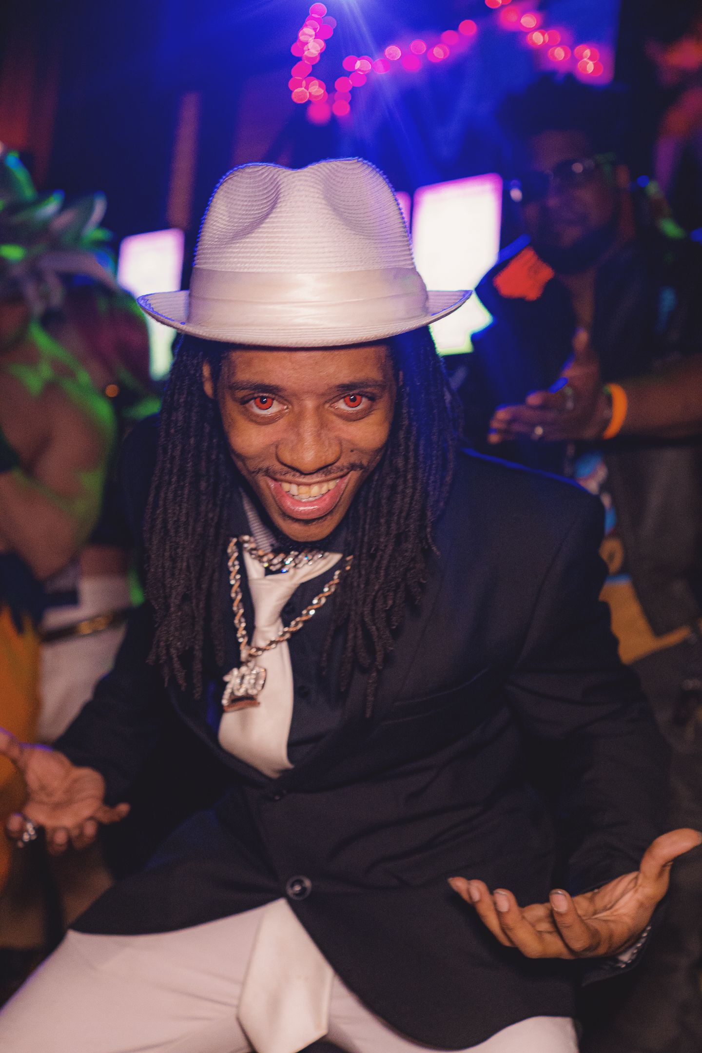 Smiling man with dreadlocks wearing a white fedora and black blazer with chain necklaces at a vibrant nightclub event.