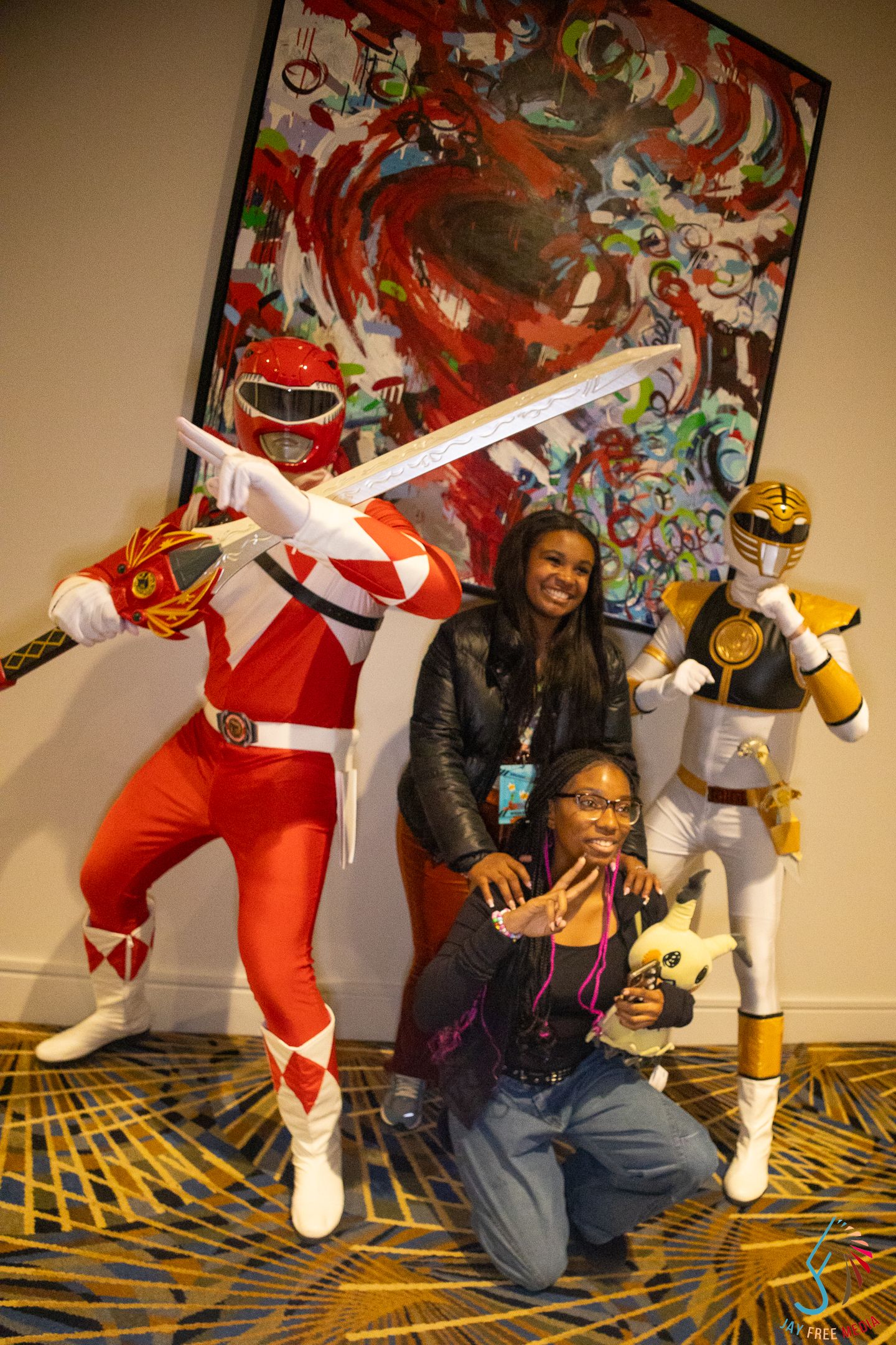 Fan poses with Red and White Power Rangers cosplayers holding a Pikachu plush at a convention.