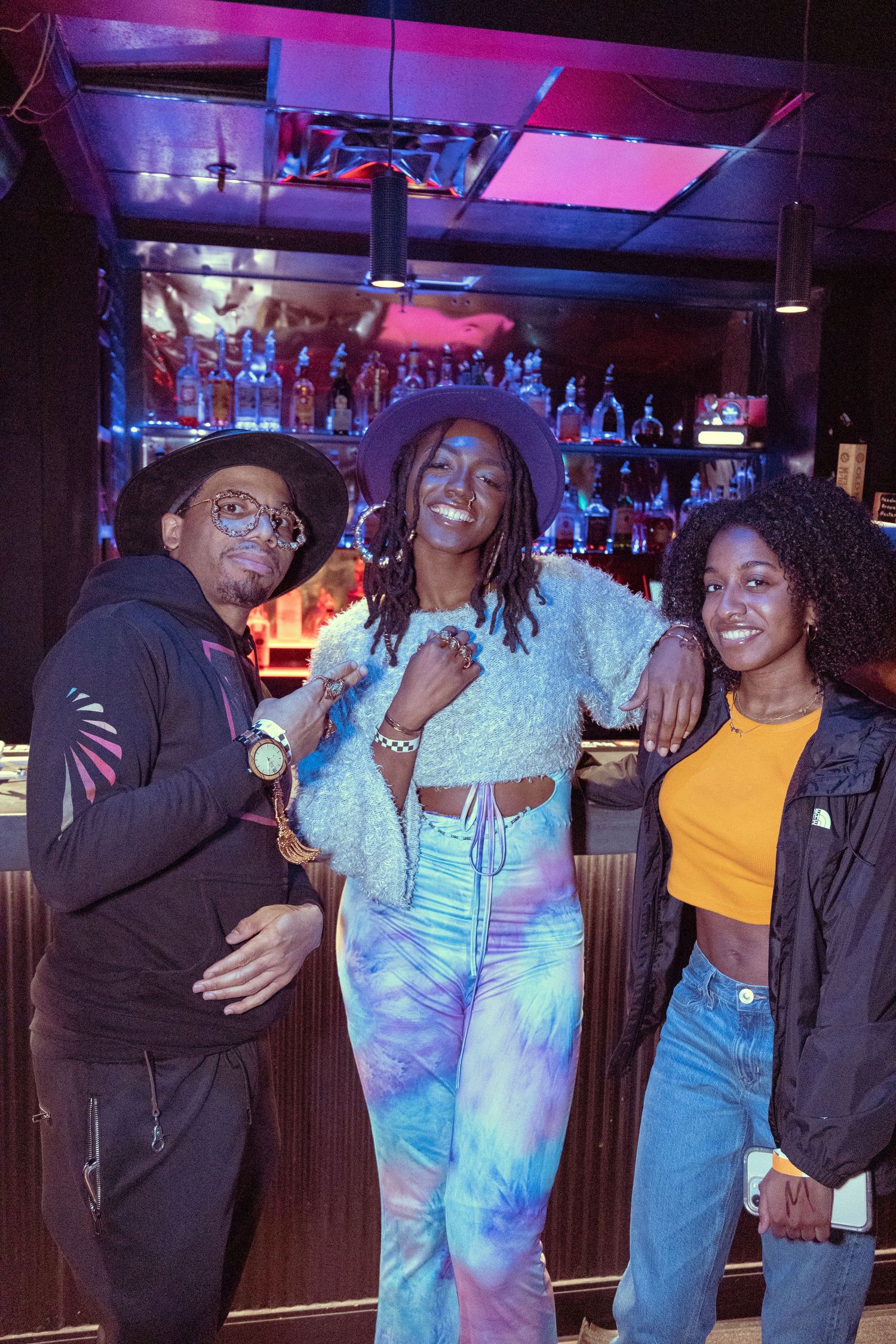 Three friends posing together at a bar, with a woman in tie-dye outfit flanked by two smiling companions.