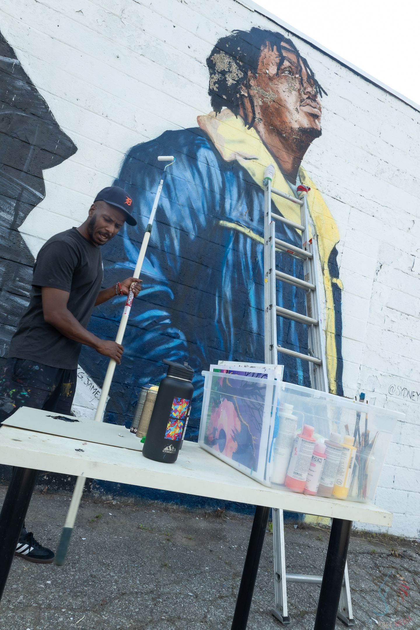 Artist stands at work table in front of large colorful mural painted on brick wall outdoors.