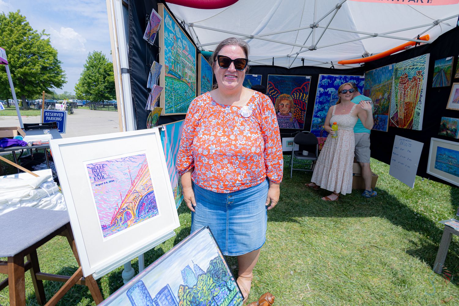Woman in floral top browses colorful artwork displays at an outdoor art fair booth.