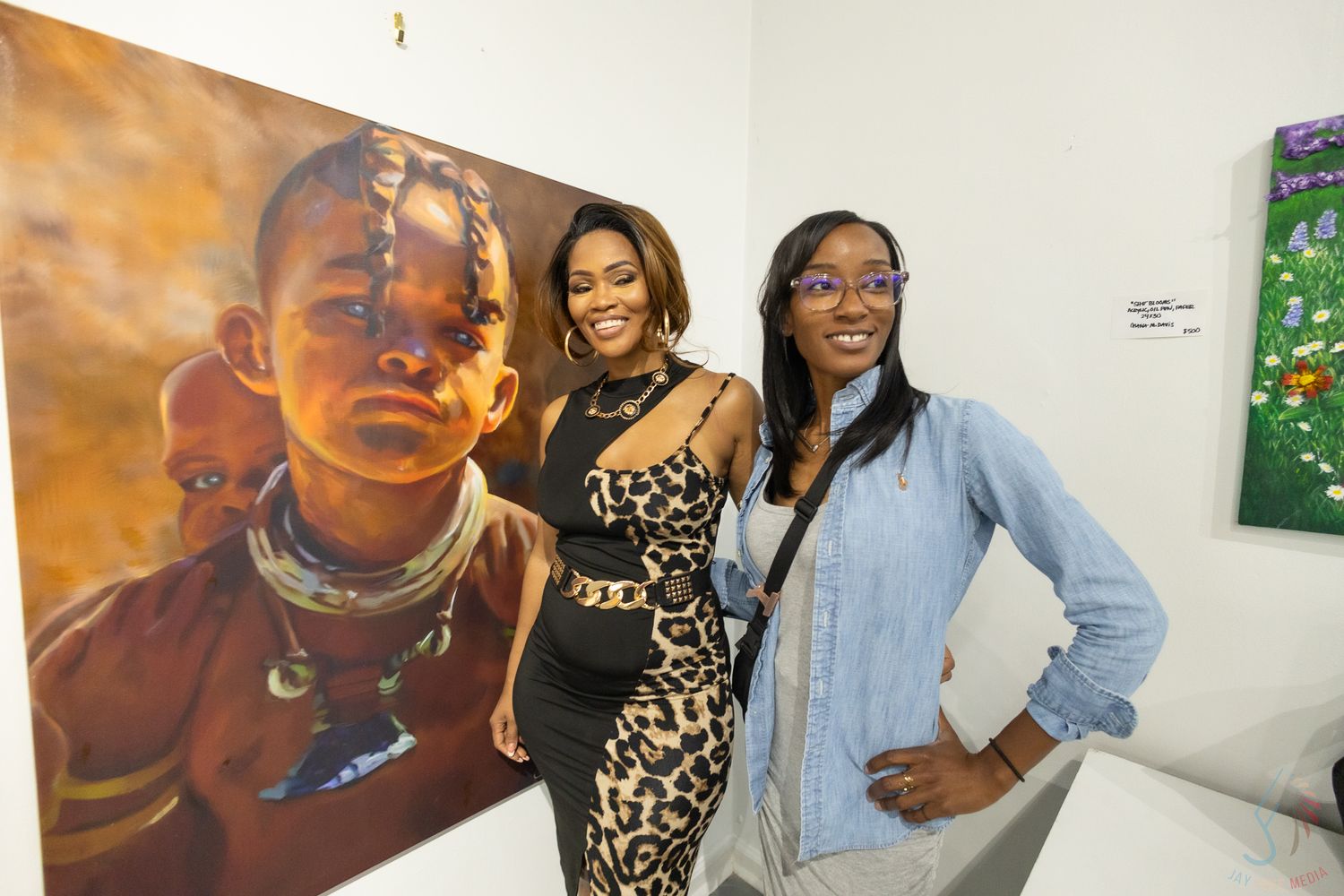 Two women posing beside a large portrait painting of an African child at an art gallery exhibition.