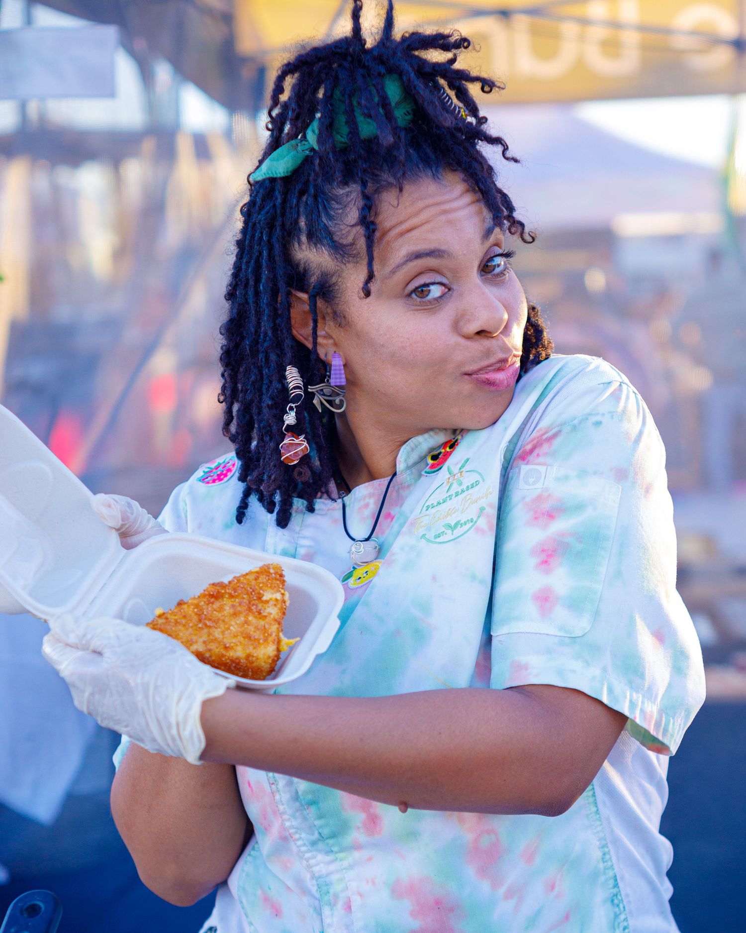 Woman with colorful locs and tie-dye shirt holds food sample at outdoor festival booth.