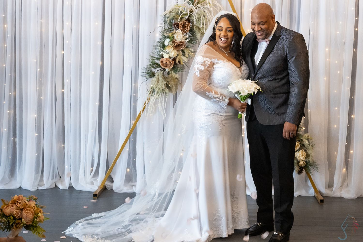 Bride and groom smiling together at altar with gold triangular arch and white drape backdrop at wedding ceremony.