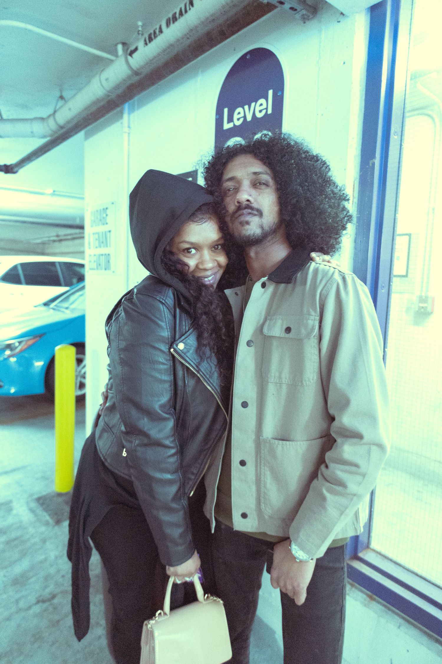Stylish couple posing in a parking garage, woman in leather jacket with hat, man in tan jacket with curly hair.