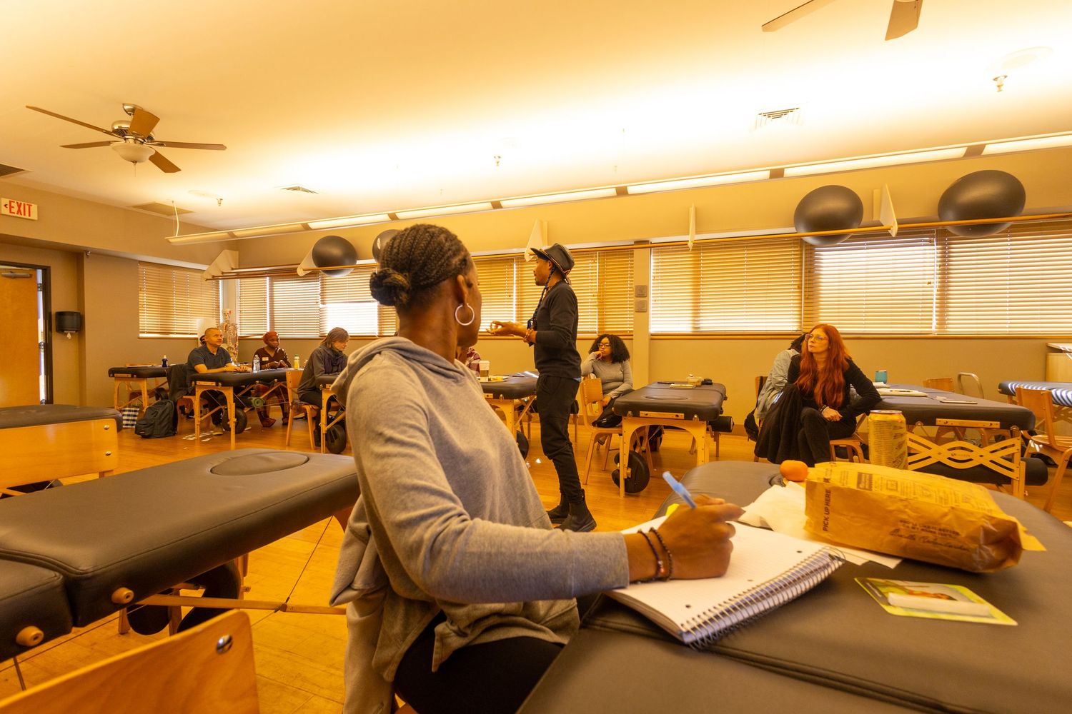 Students in a massage therapy classroom taking notes near treatment tables in a warmly lit training room.