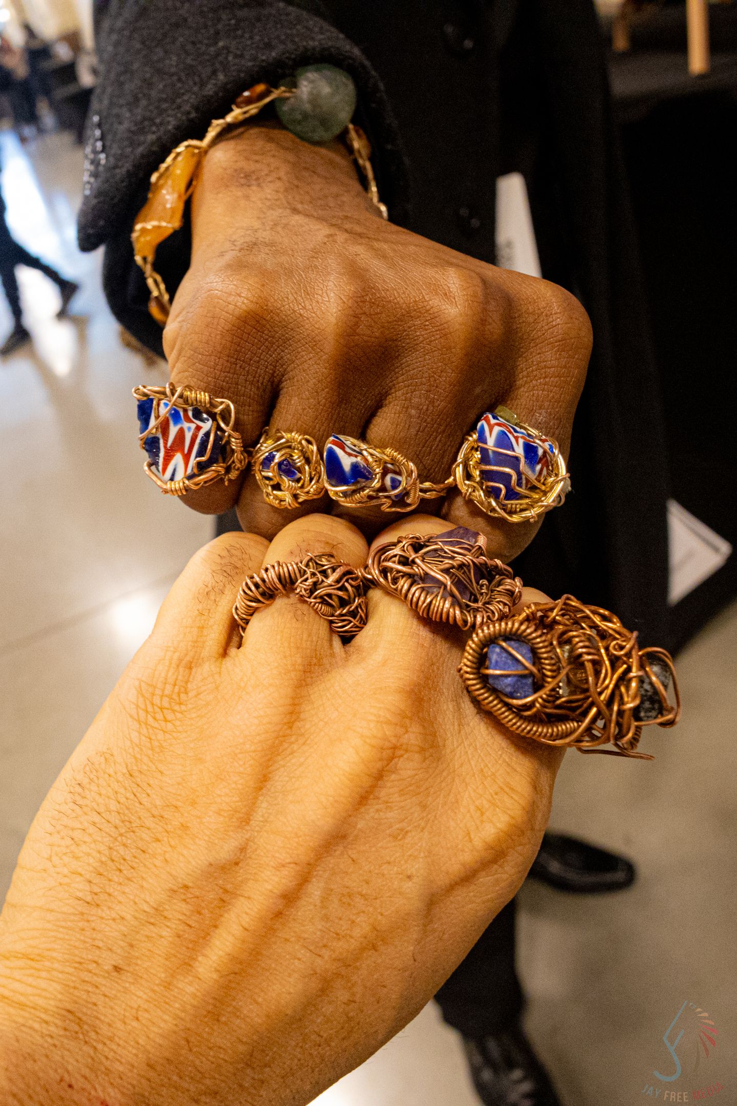 Two hands adorned with copper wire-wrapped blue gemstone rings and matching bracelets at a jewelry exhibition.