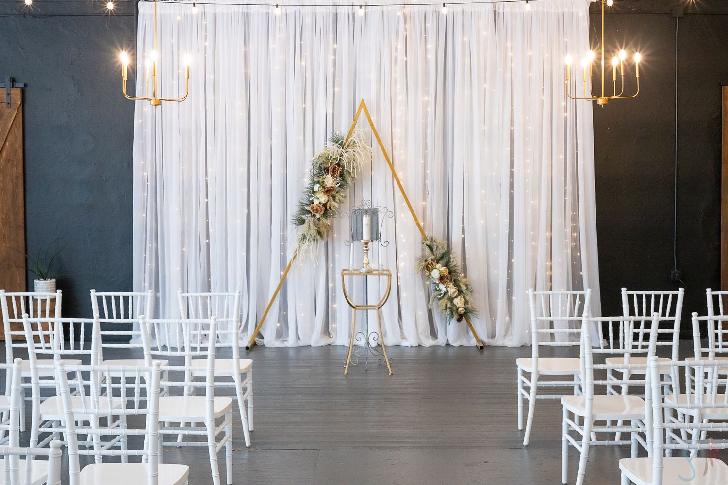 Elegant wedding ceremony setup with white draping, gold triangular arch, floral arrangements, and white Chiavari chairs.
