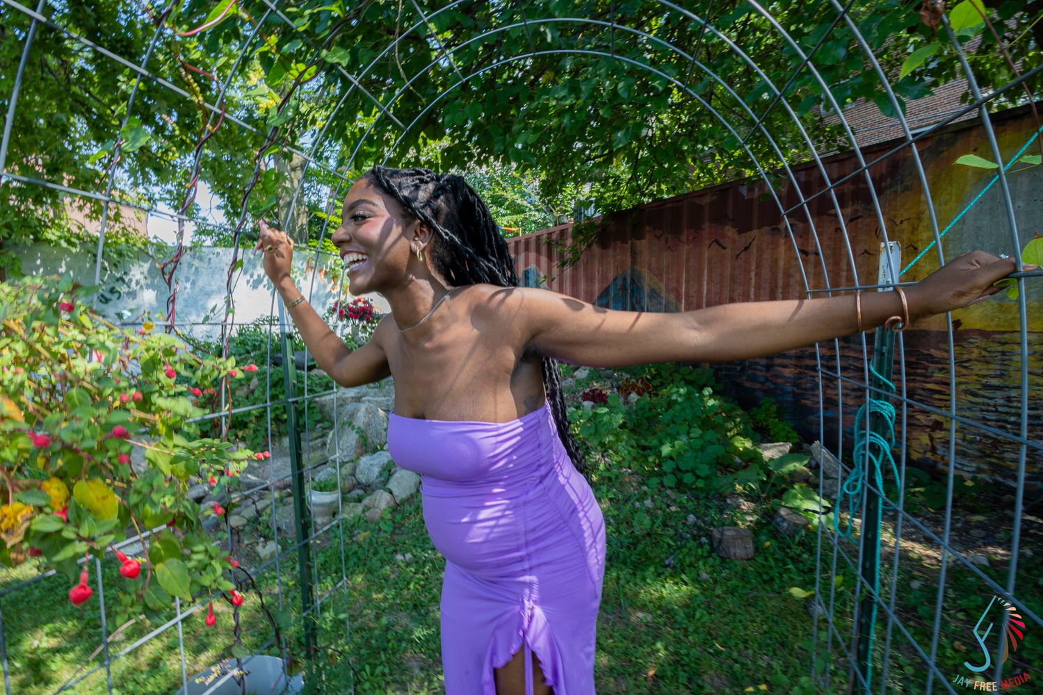 Woman in purple dress posing under green garden arch surrounded by lush plants and flowers on sunny day.