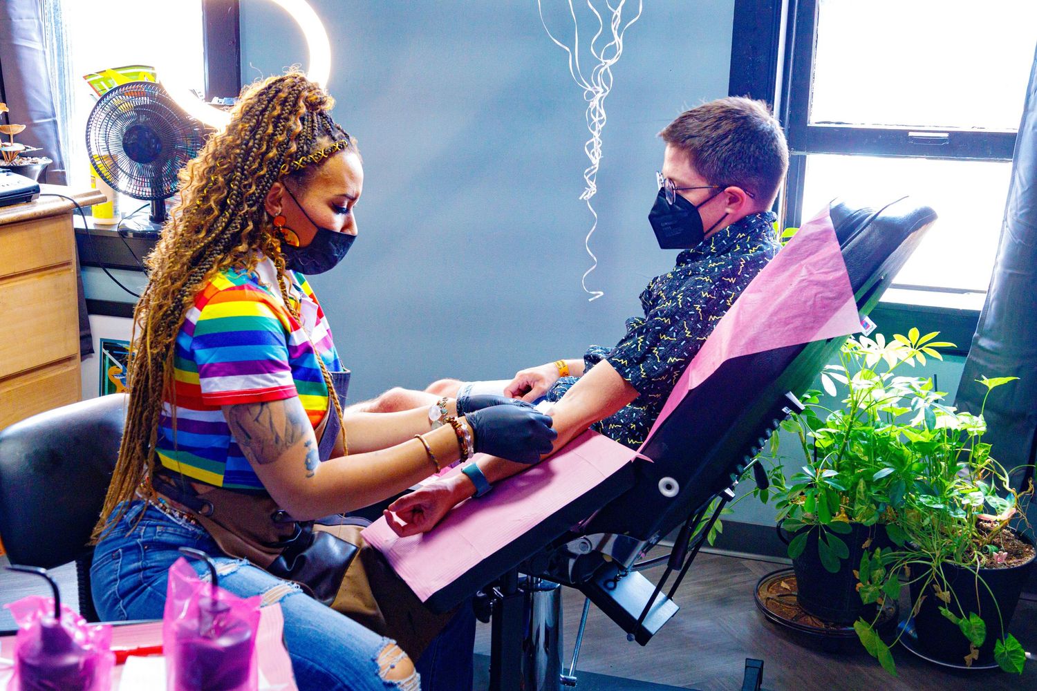 Nail technician in rainbow shirt applies manicure to client wearing mask in cozy salon setting.