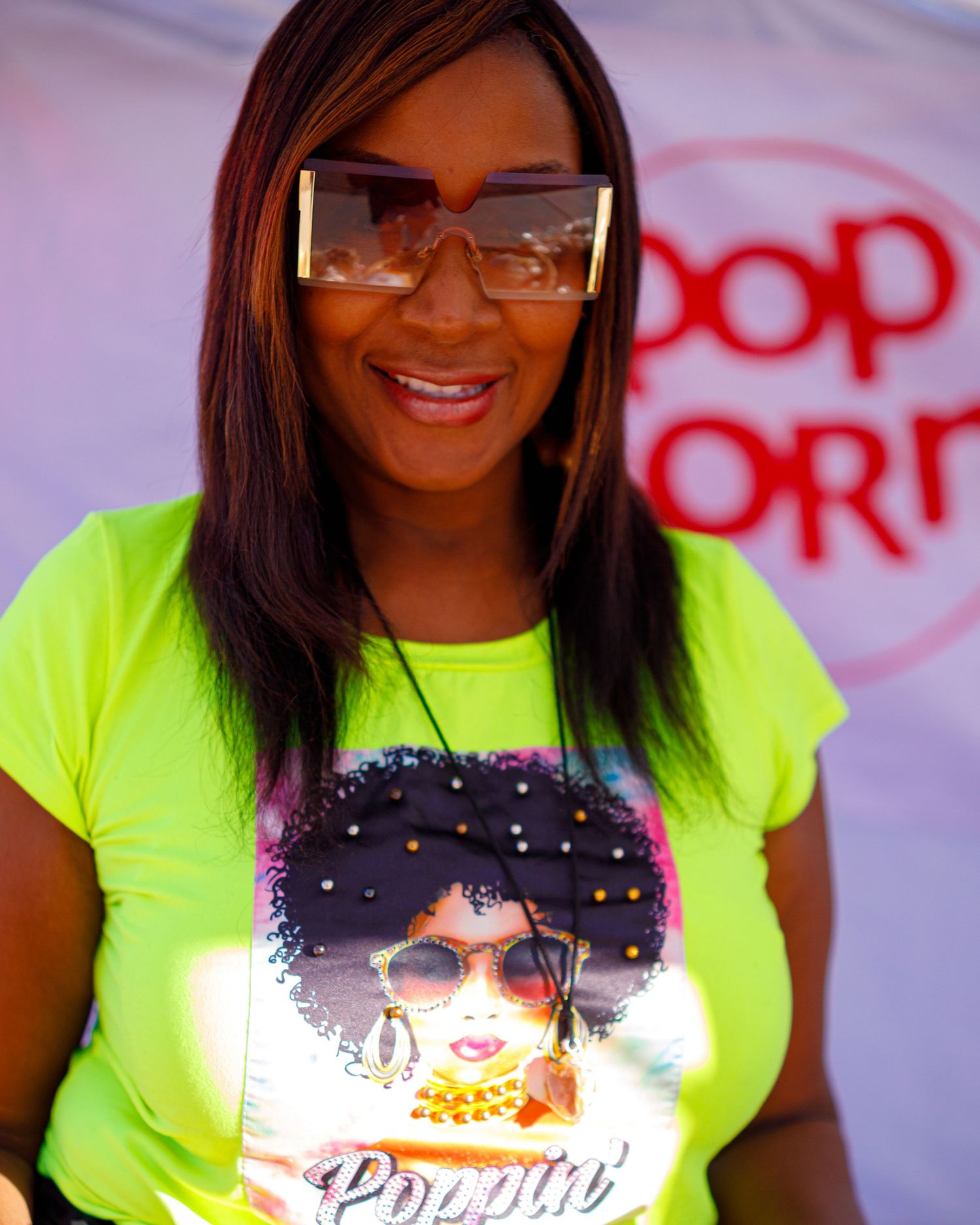 Woman in neon yellow graphic tee and oversized sunglasses smiling at outdoor event.