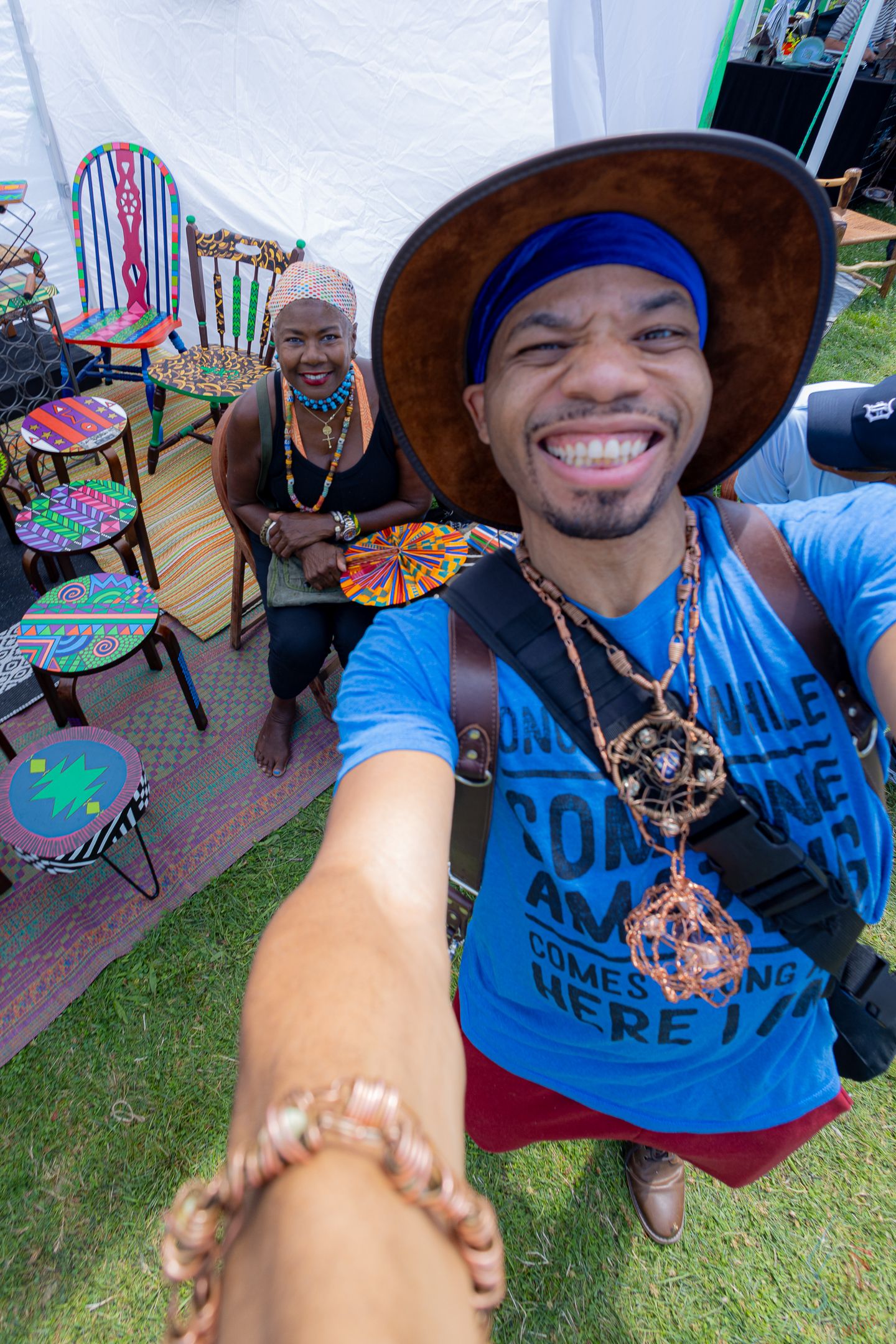 Young man in blue shirt and brown hat takes selfie with smiling woman beside colorful painted furniture at art fair.
