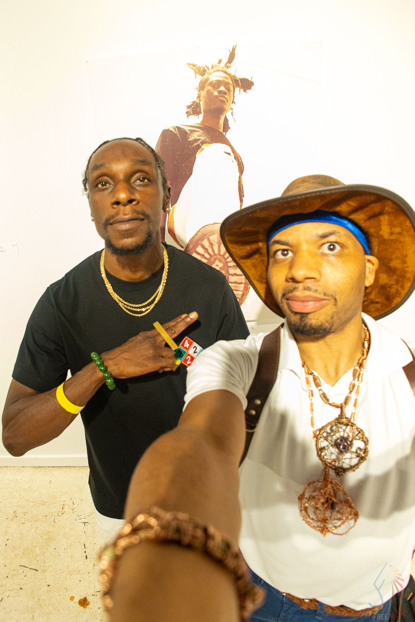 Two men posing for a selfie in an art gallery, one wearing a hat and large pendant necklace, the other in a black tee.
