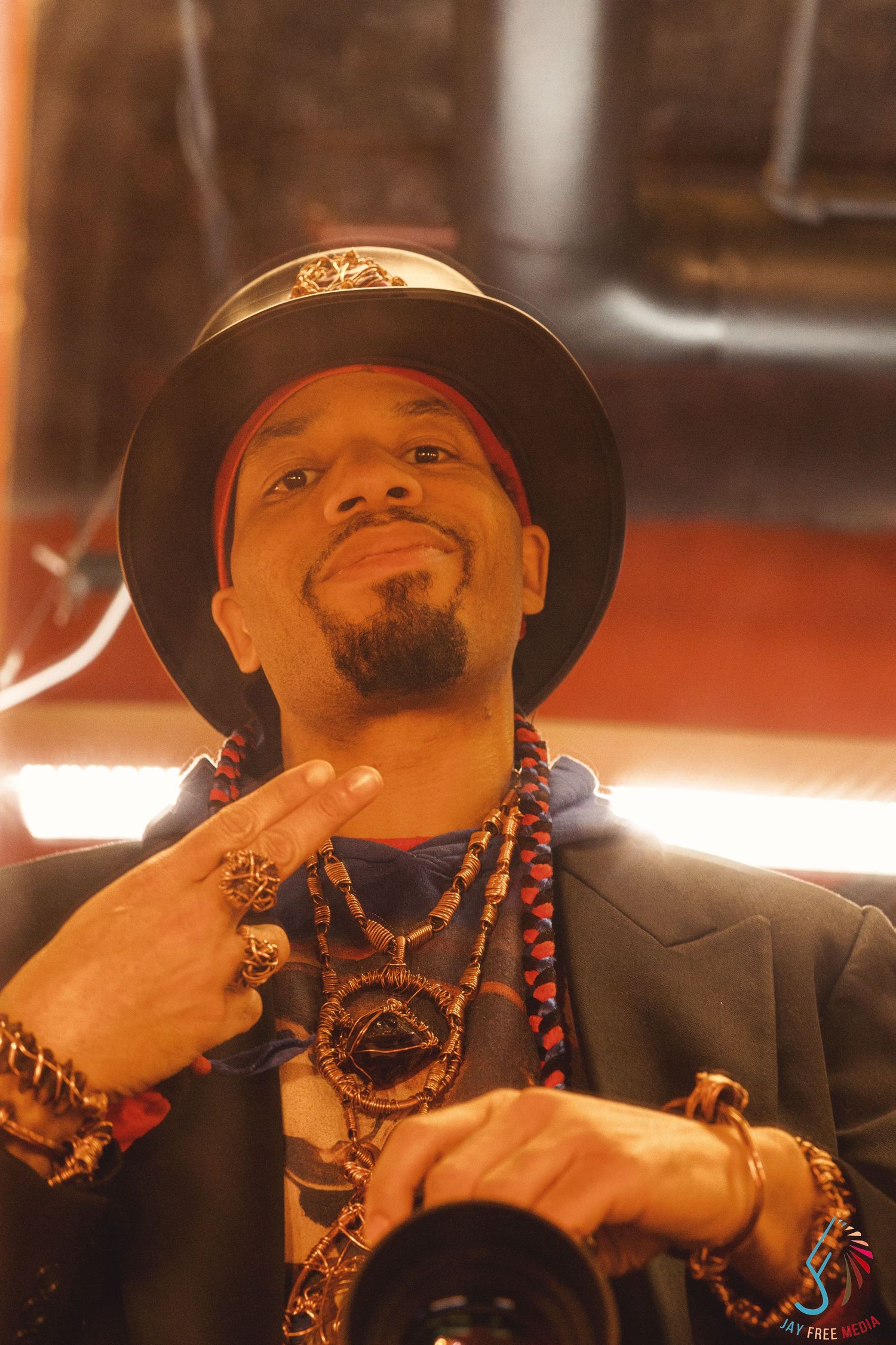 A stylish man wearing a black hat and layered gold jewelry poses with a hand gesture indoors.