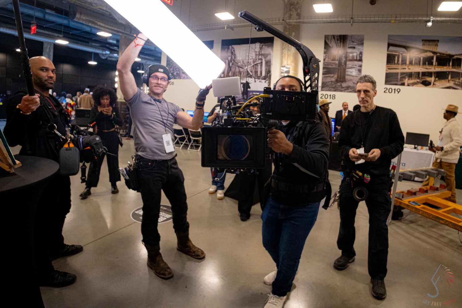 Film crew members carry professional camera equipment through an indoor event space during a live production shoot.