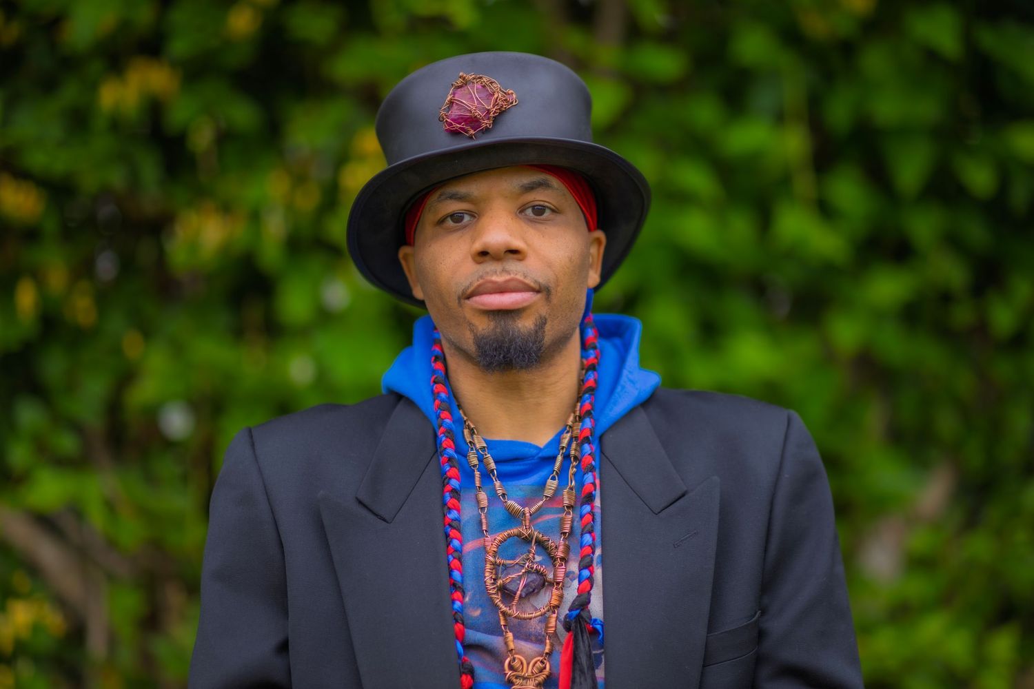 A stylish man wearing a black top hat, blue hoodie, and colorful beaded necklaces poses outdoors.