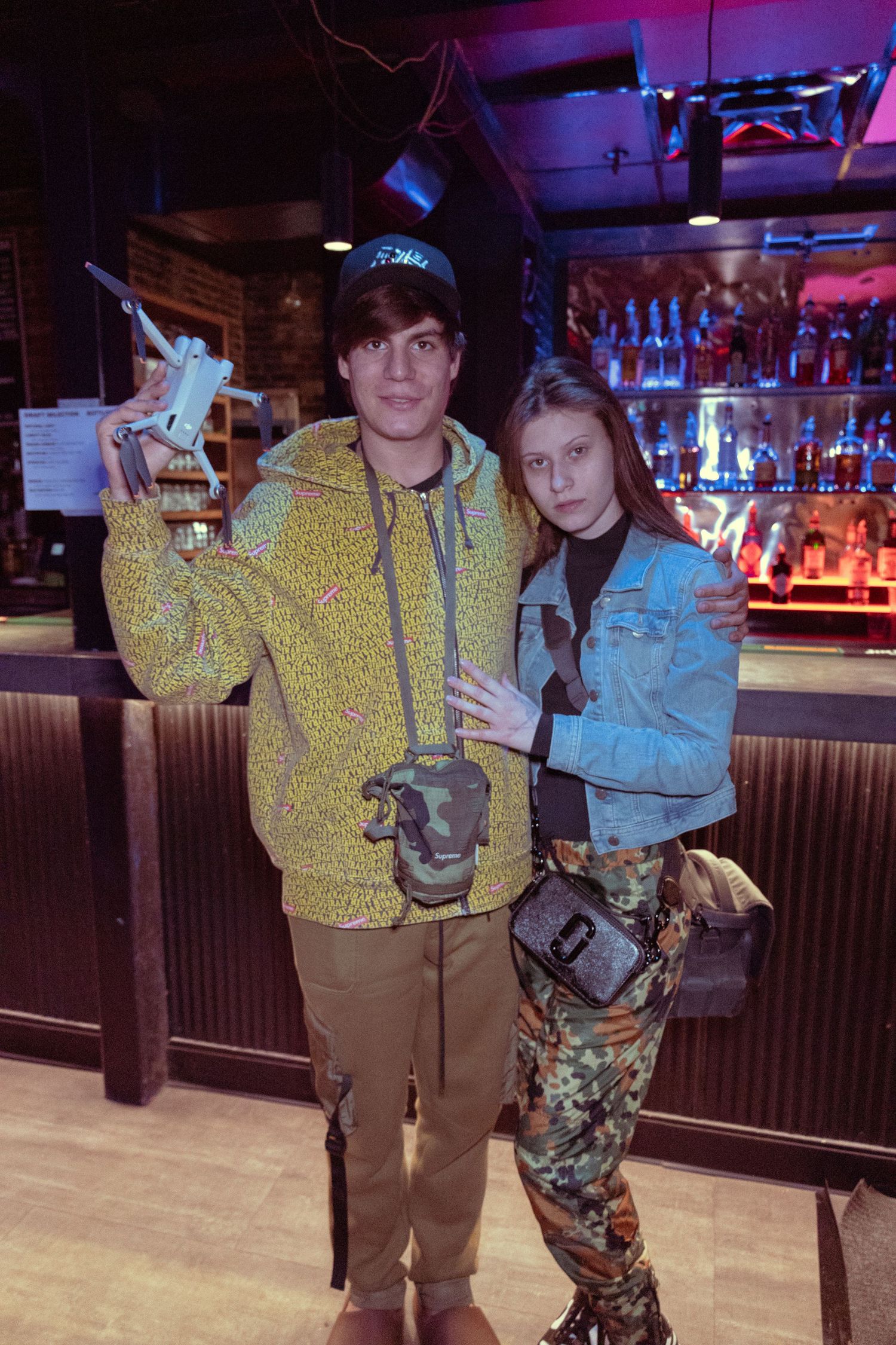 Young couple posing together at a bar, man in yellow jacket holding trophy, woman in denim jacket and camo pants.