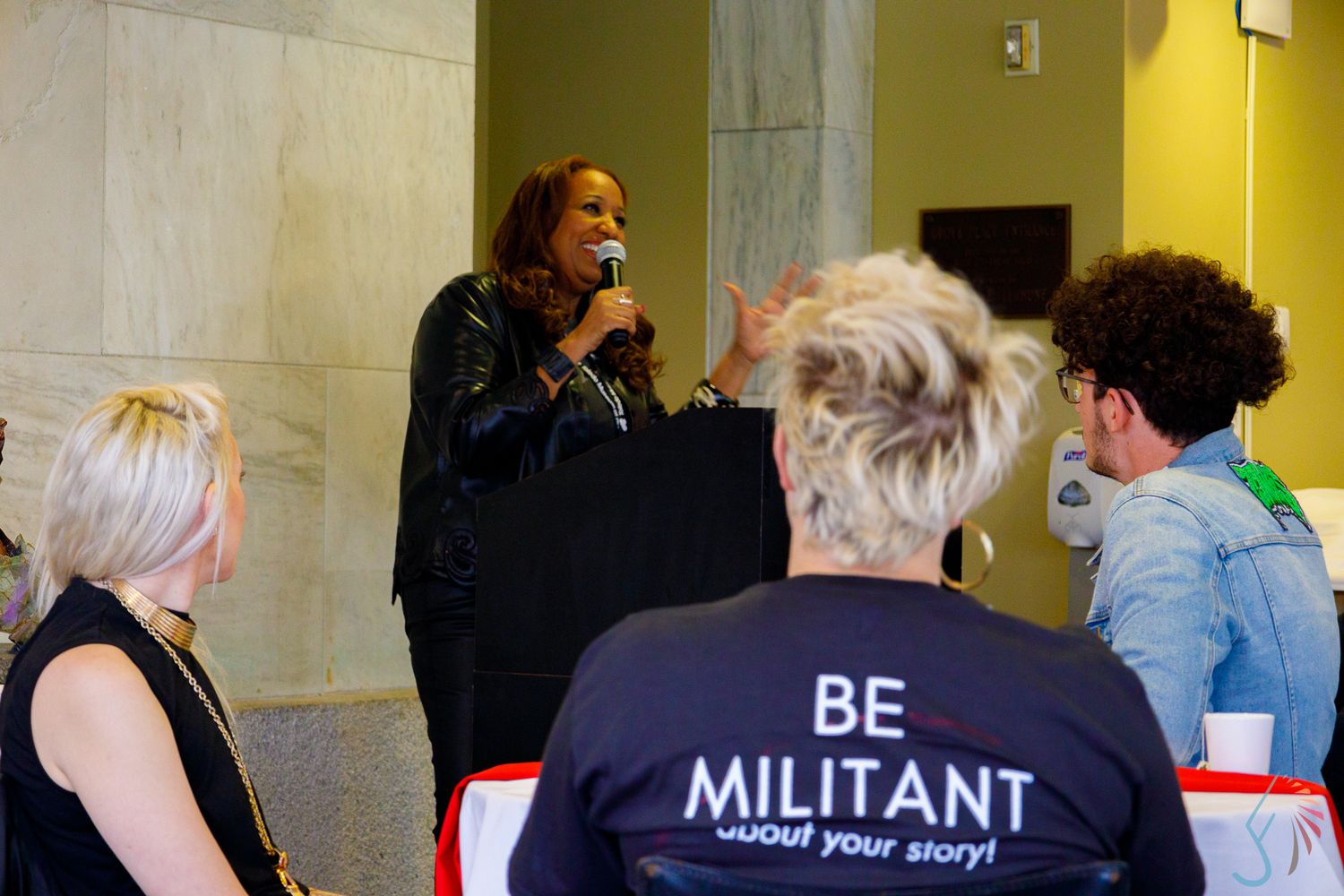 Woman speaks at podium to seated audience, one wearing a 'Be Militant About Your Story' shirt, at an indoor event.