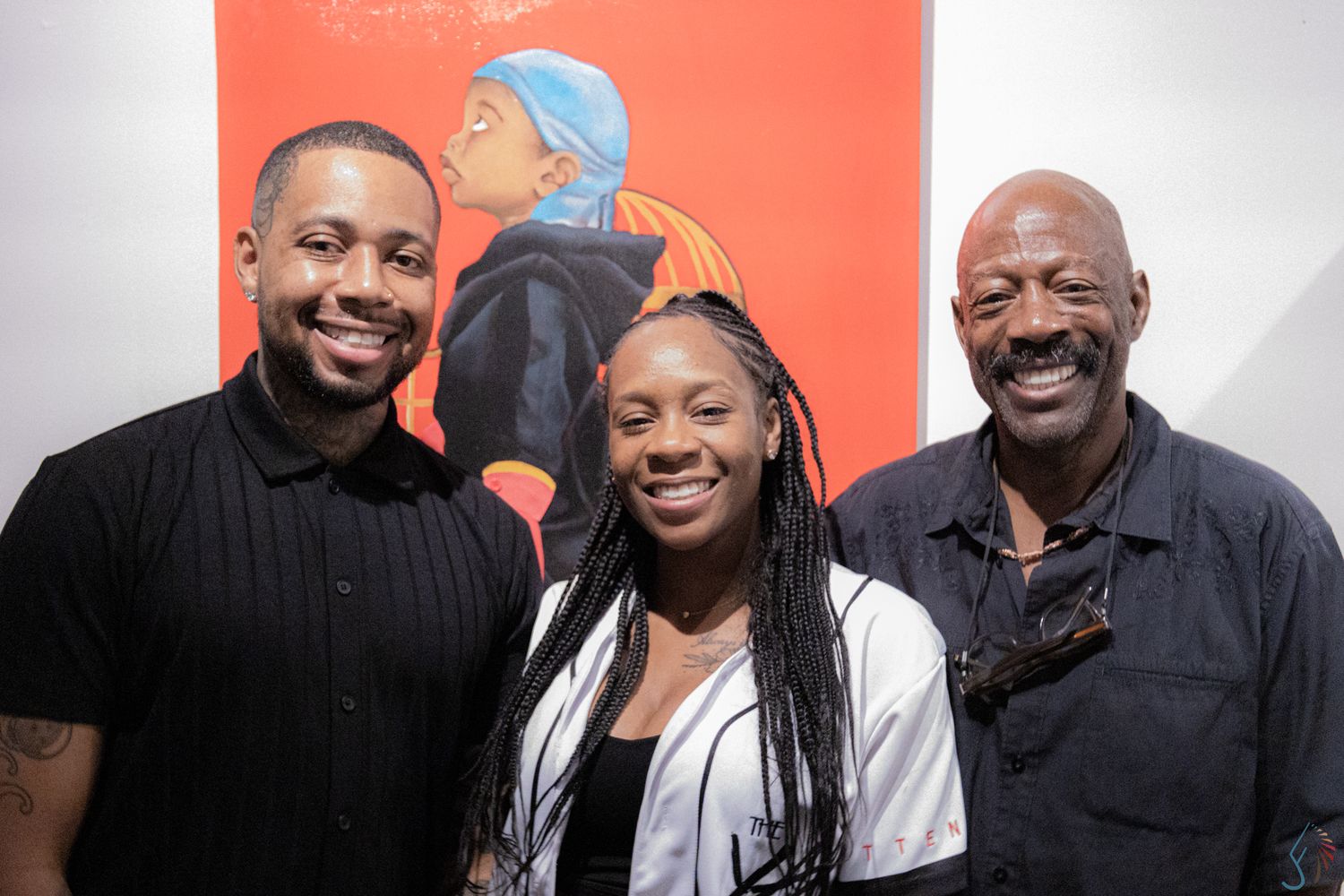 Three people smiling in front of a vibrant red portrait painting at an art gallery exhibition.