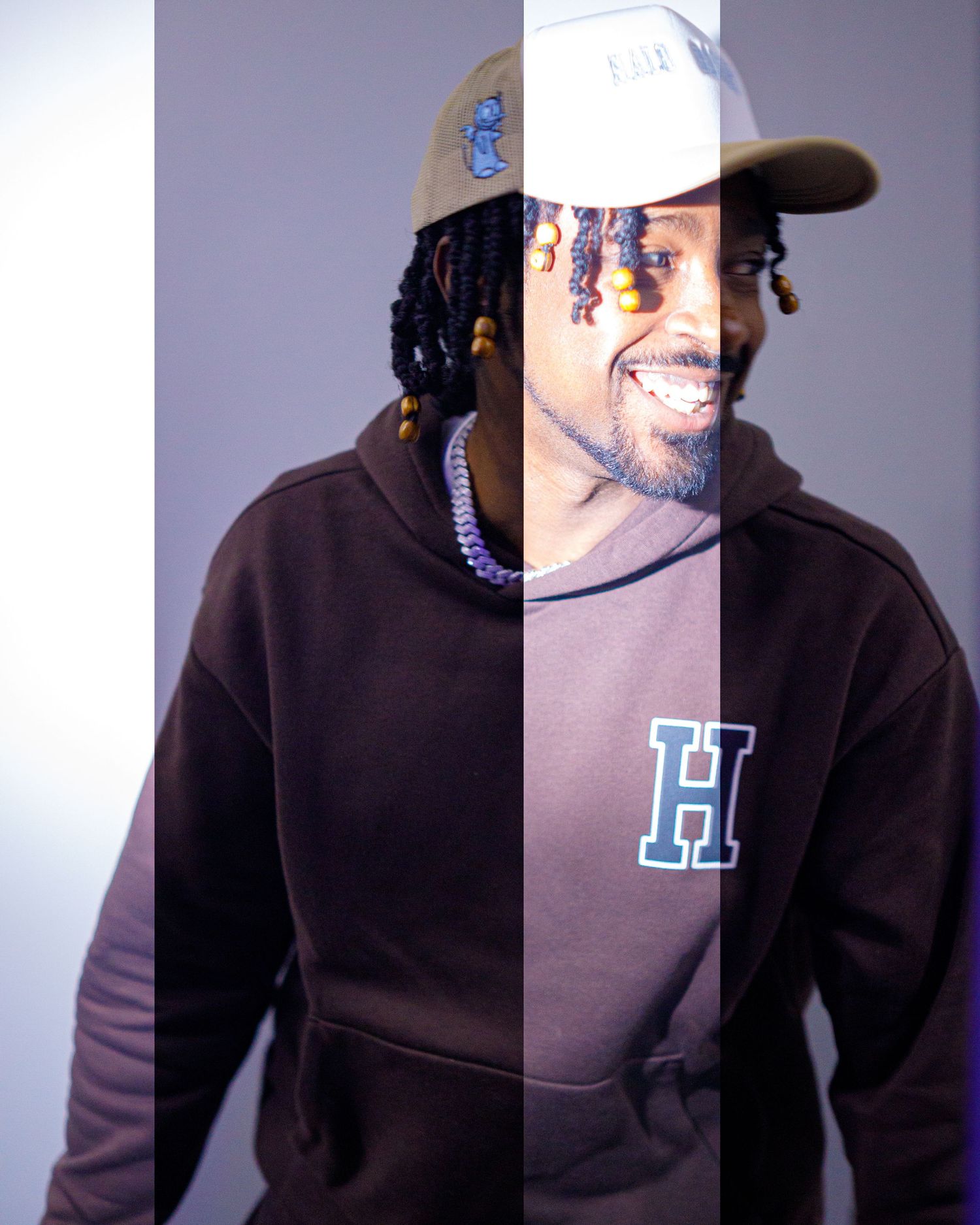Smiling man with braids wearing a brown HUF hoodie and tan cap in a split-tone portrait photo.