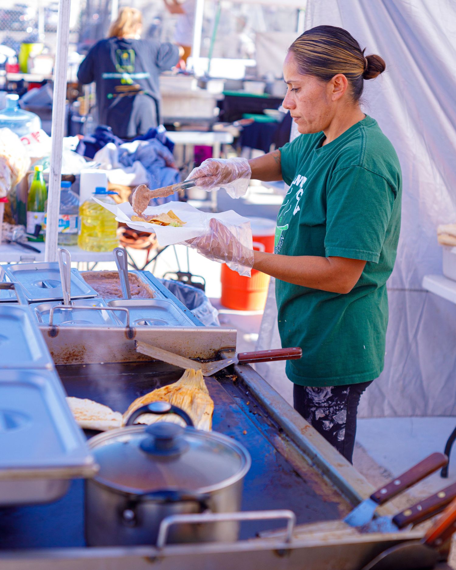Woman in green shirt cooking on a flat top grill at an outdoor food market stall.